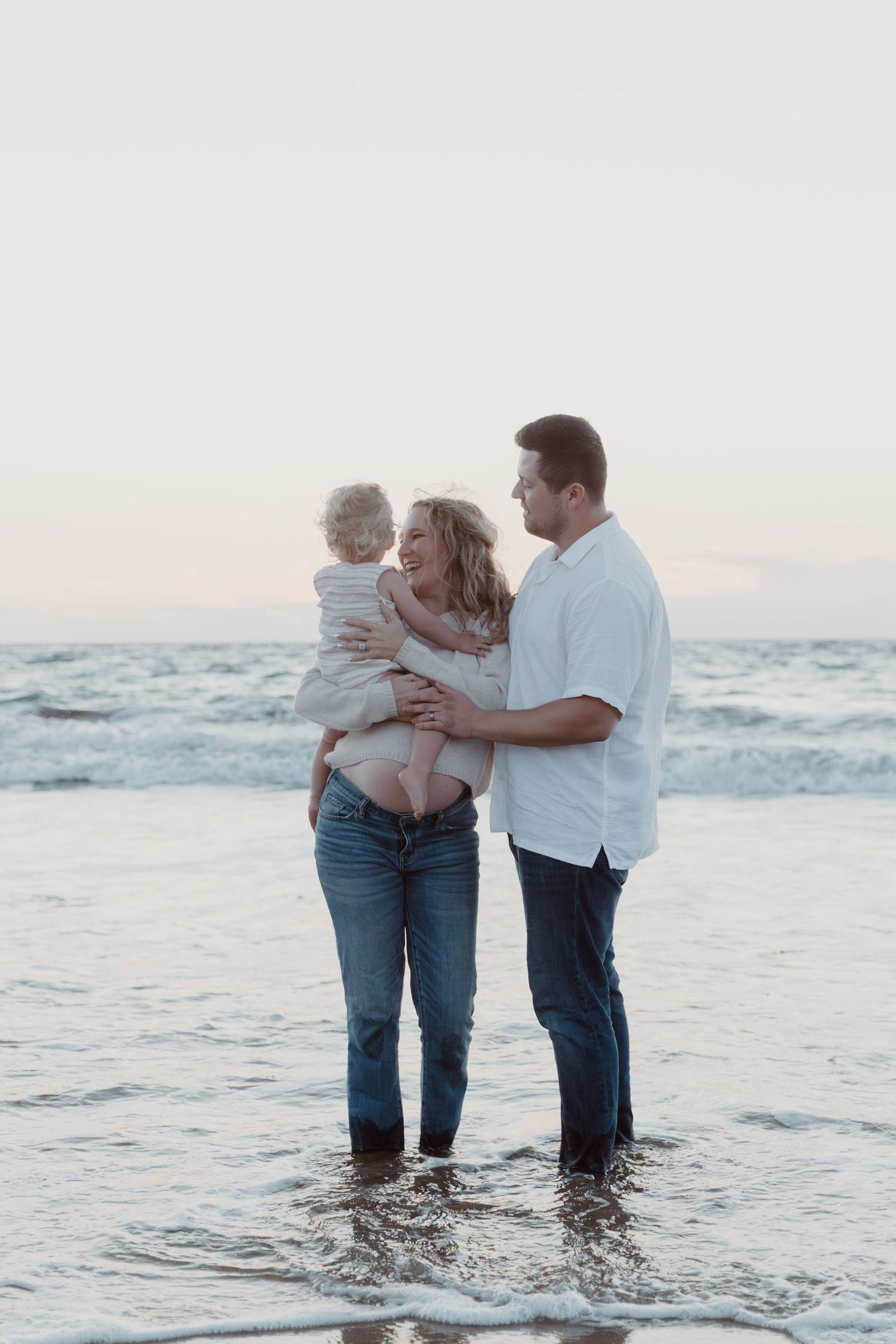 Family standing together in shallow ocean waves during a beach photoshoot at sunset.