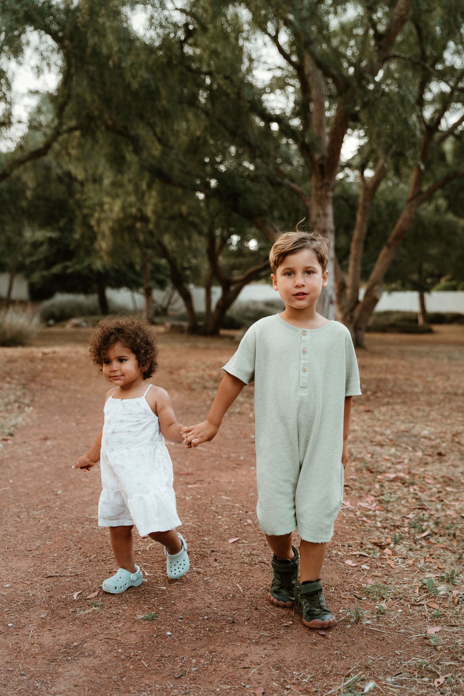 Two children walking together on dirt path surrounded by trees.