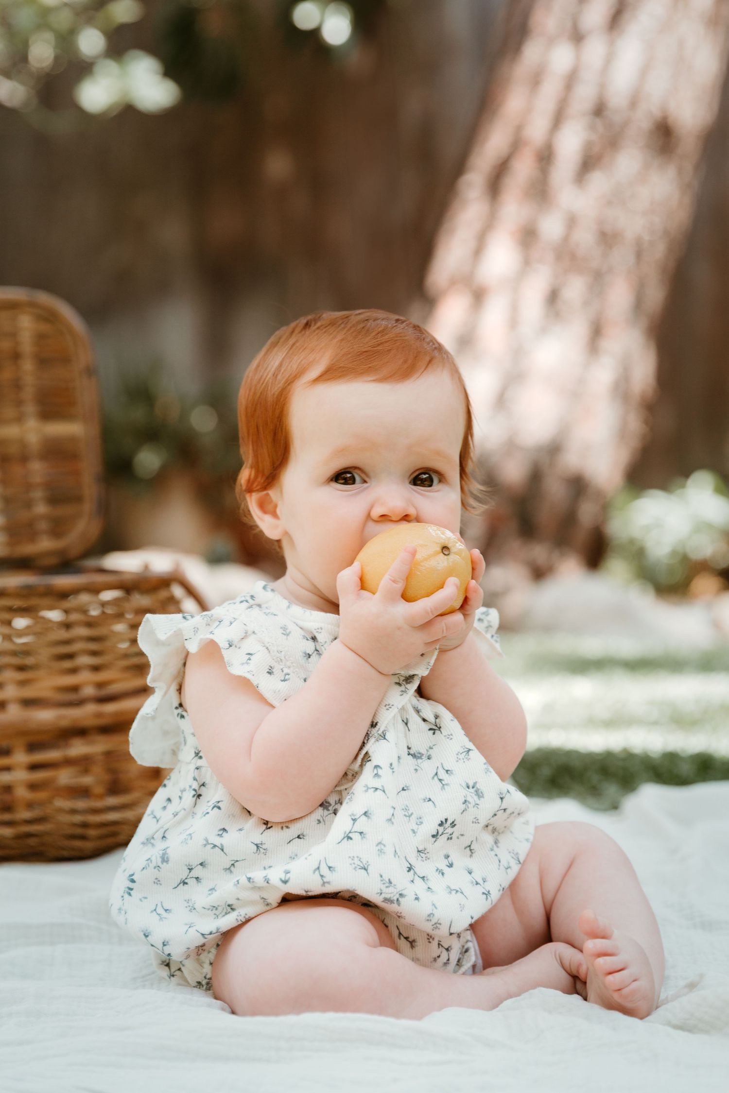 Baby in floral dress lying on white blanket near wicker basket in garden setting.