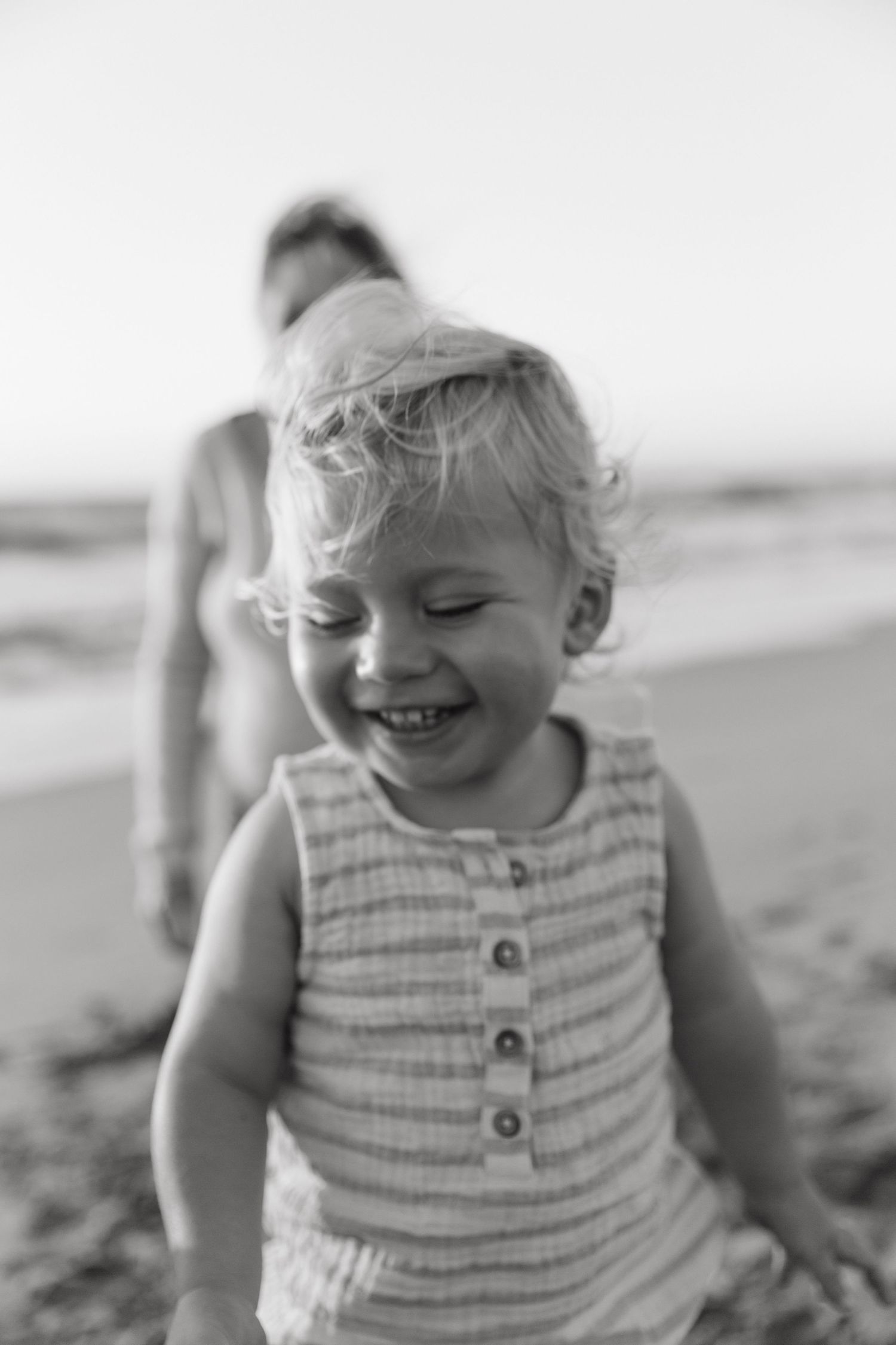 Black and white photo of a happy toddler in a striped tank top playing on the beach.