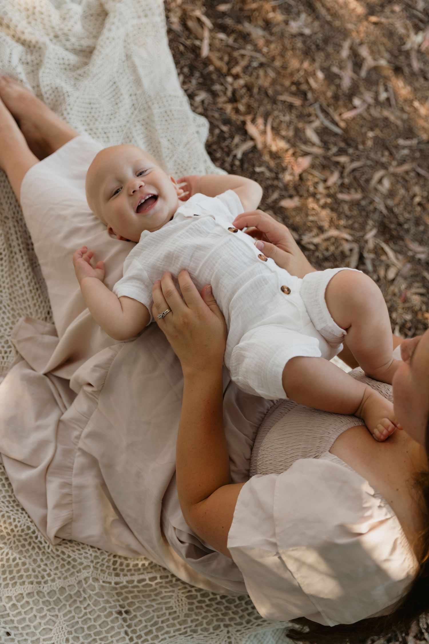 Baby in white romper resting on cream blanket in dappled sunlight.