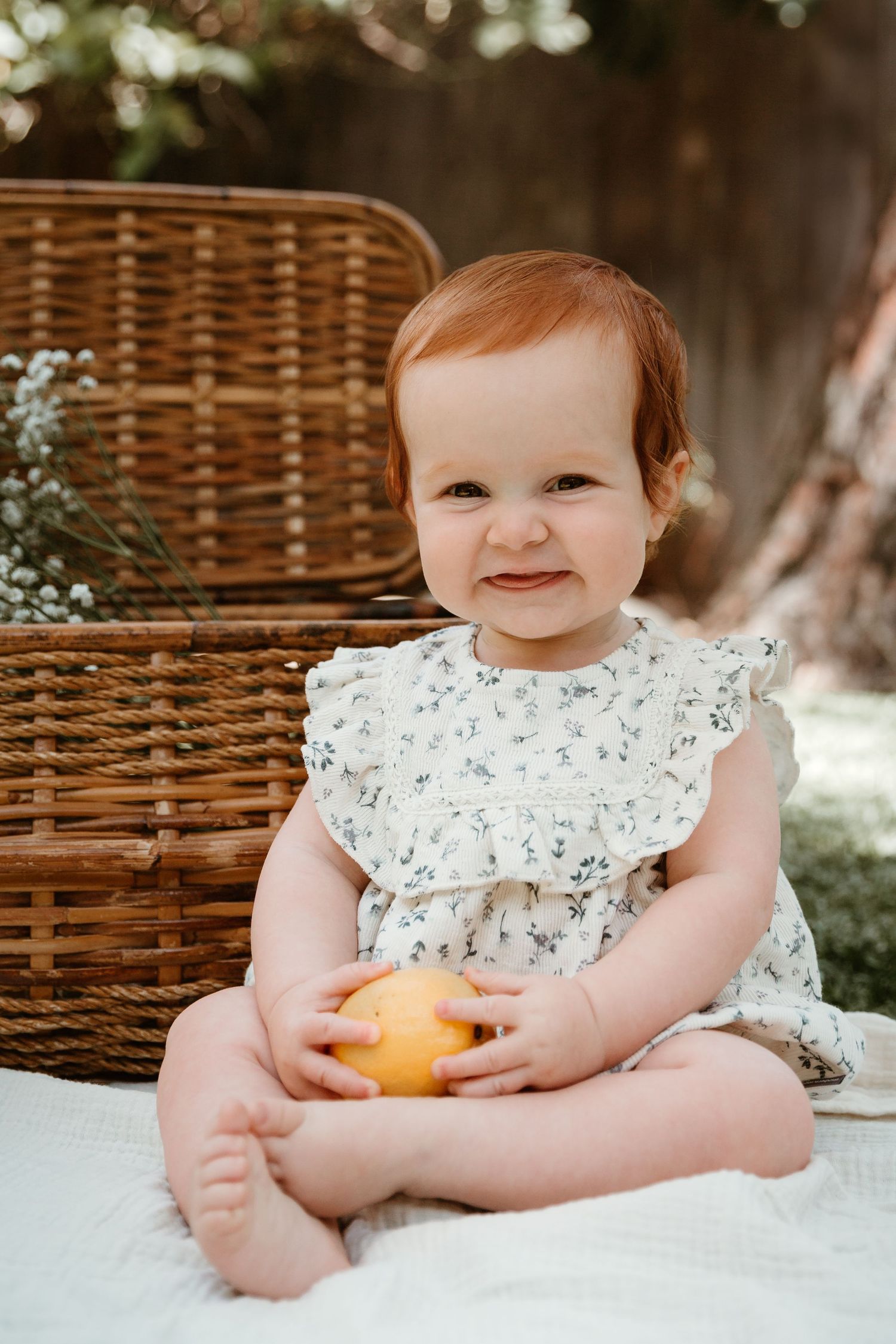 Baby in a white floral dress smiles while holding an orange on a blanket outdoors.