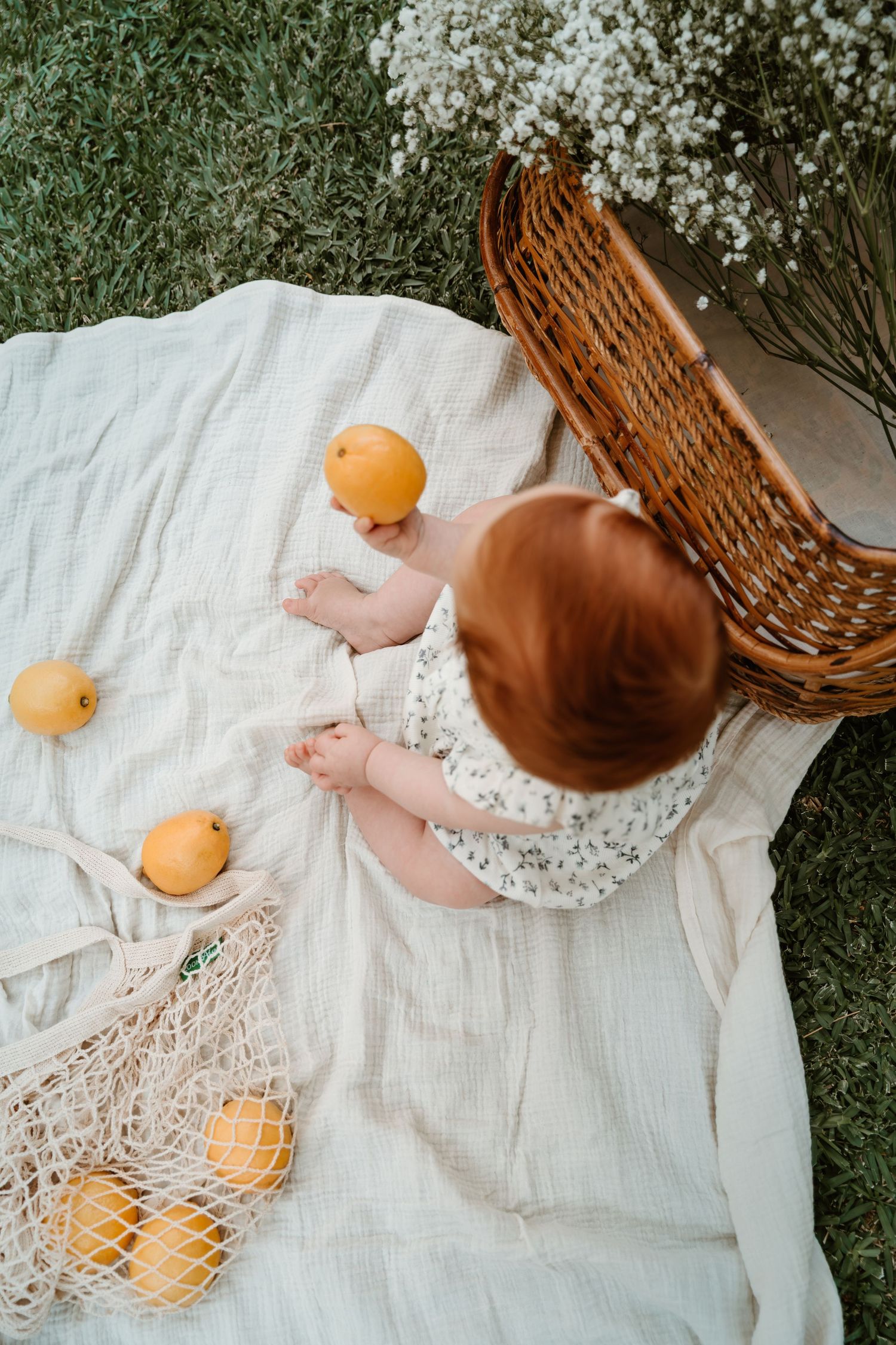 Overhead view of a baby playing with oranges on a white blanket next to a wicker basket.