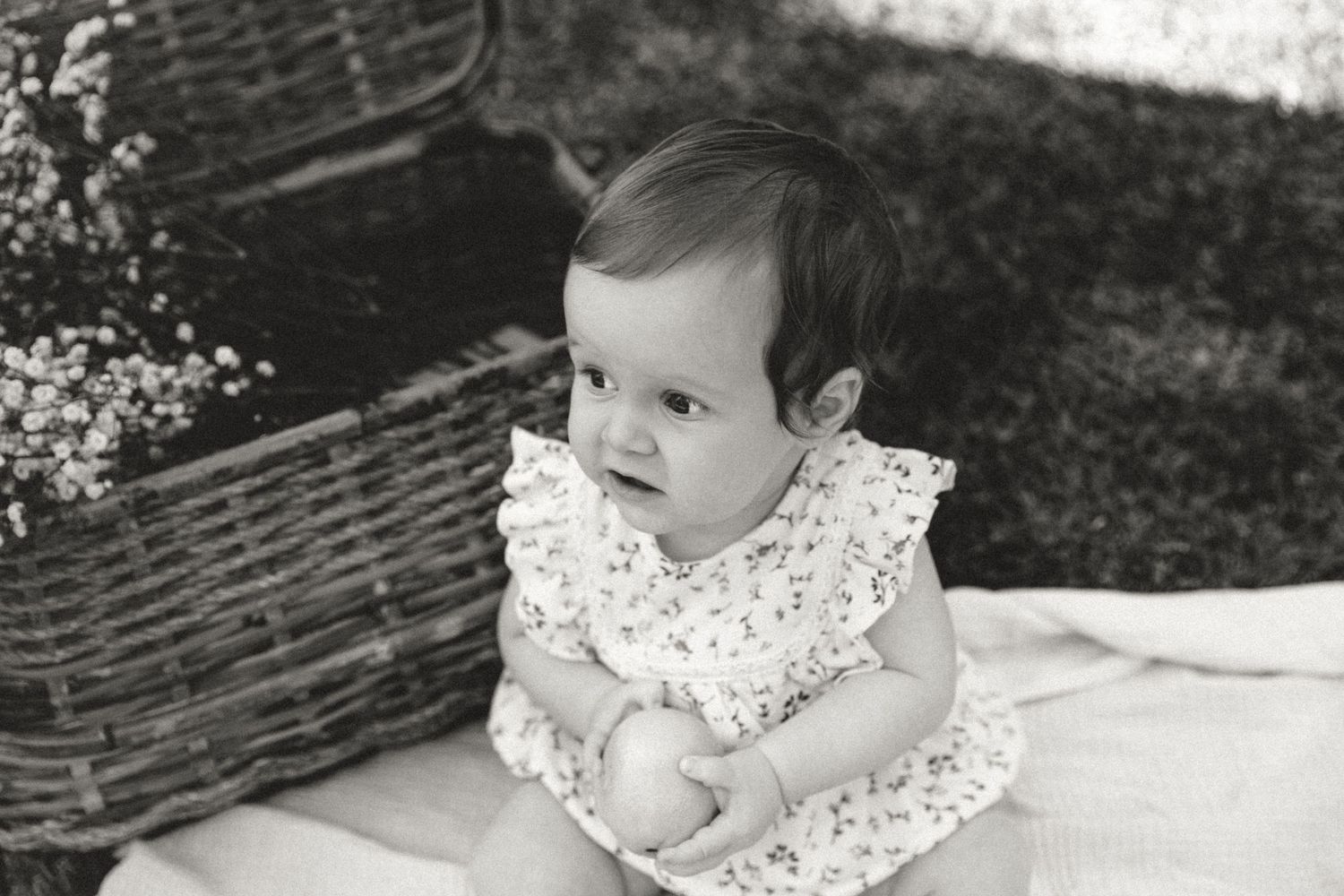 A toddler in a white dress sits among delicate dried flowers and woven baskets.
