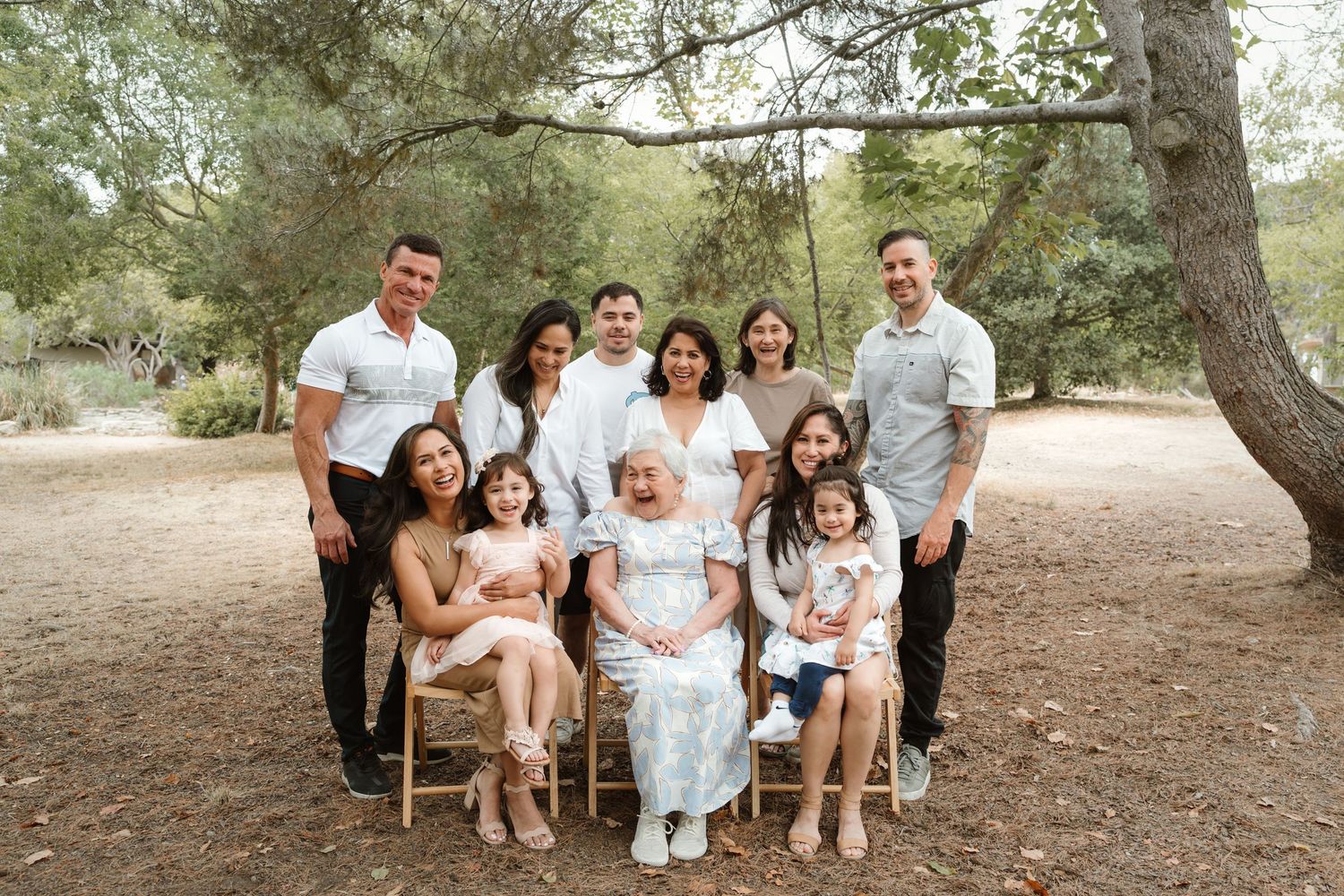 A family group poses together outdoors under shady trees in coordinated neutral-colored outfits.