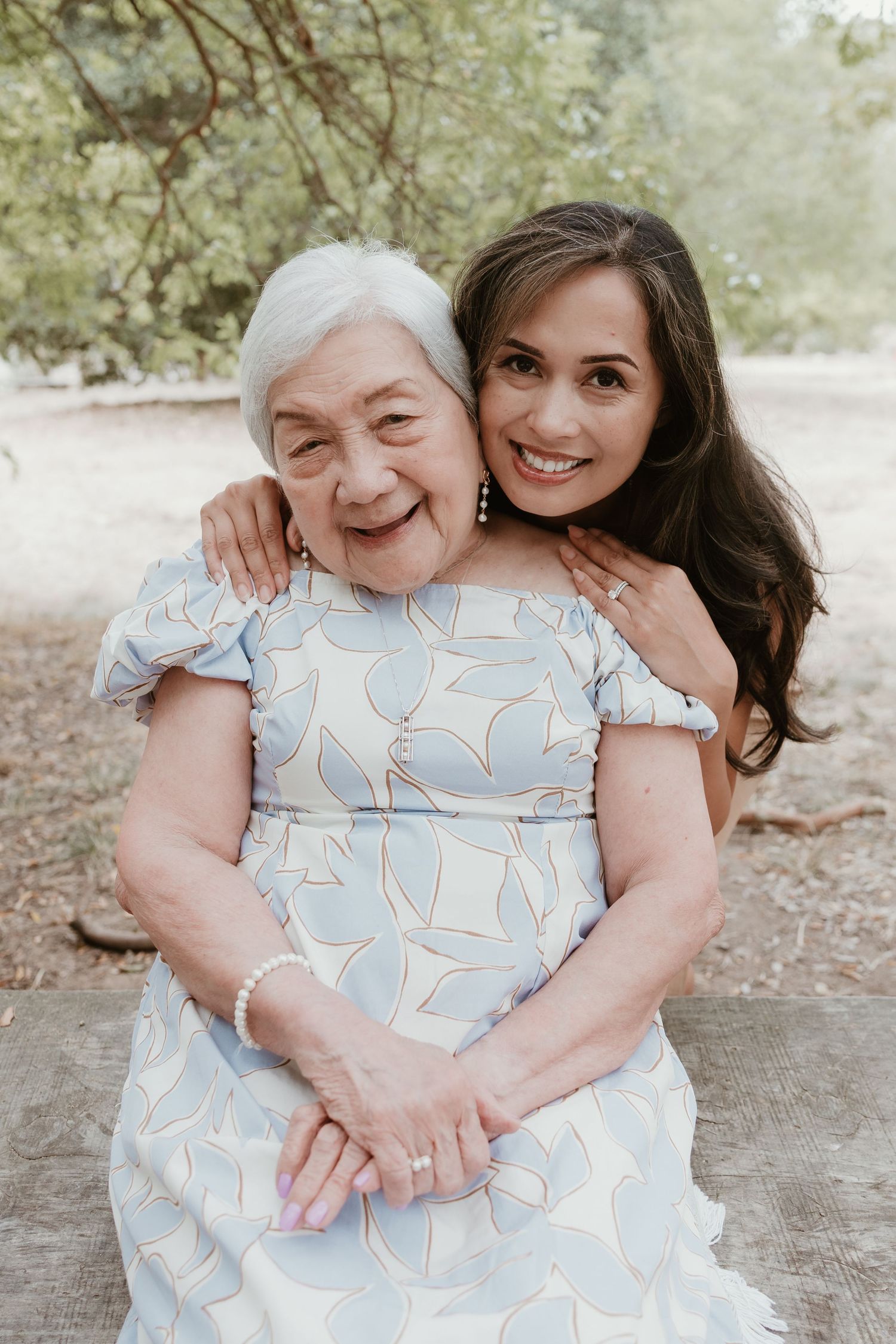 Two people share a joyful embrace while sitting outdoors in a natural setting with trees in the background.