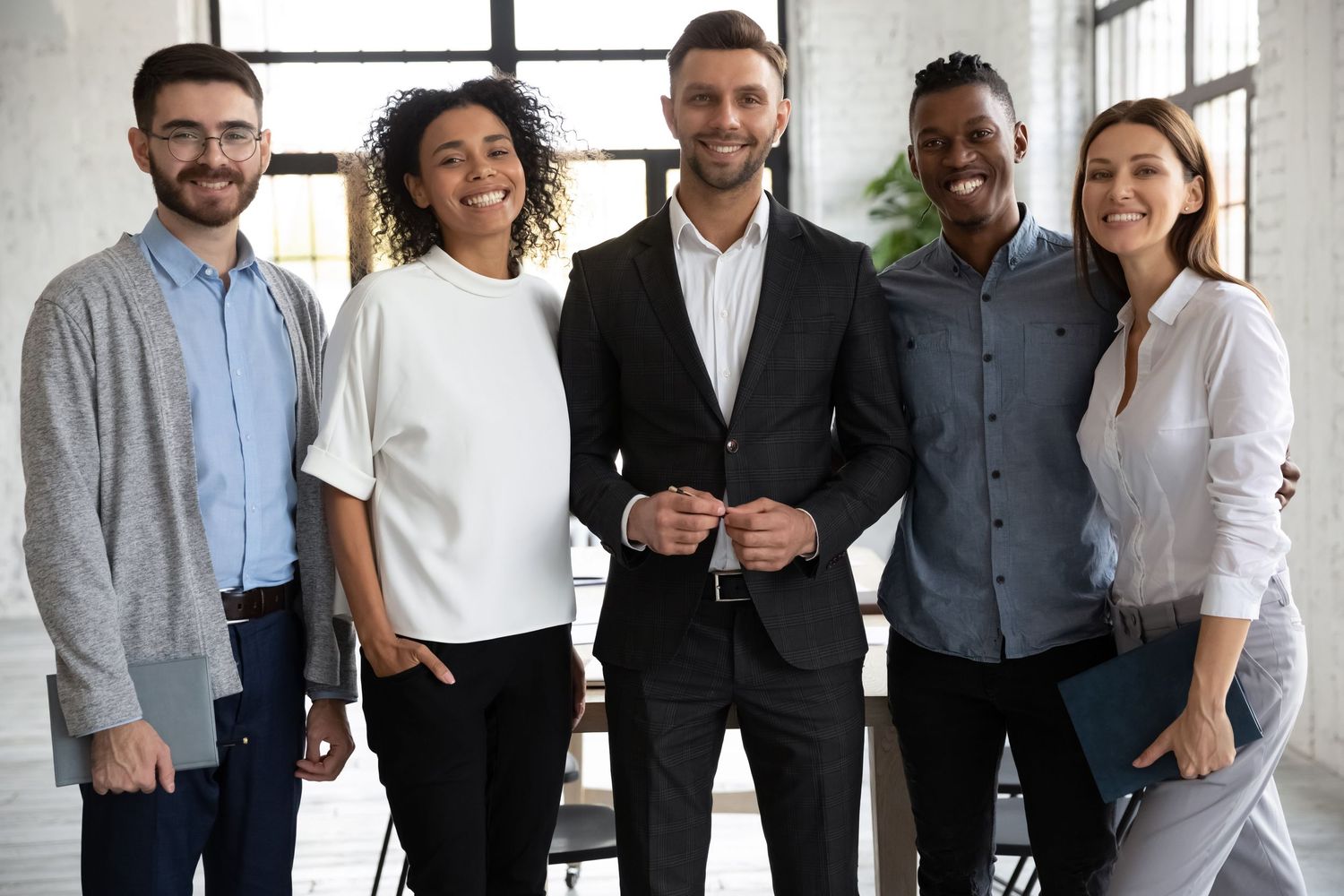 Diverse group of five smiling business professionals standing together in a modern office environment.