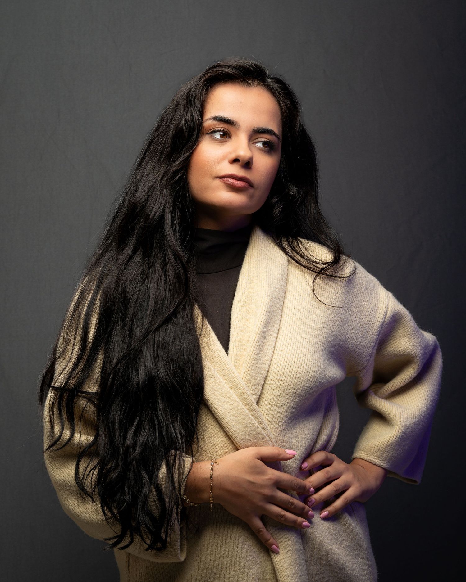 Young woman with long dark hair in beige coat posing confidently against gray studio background.