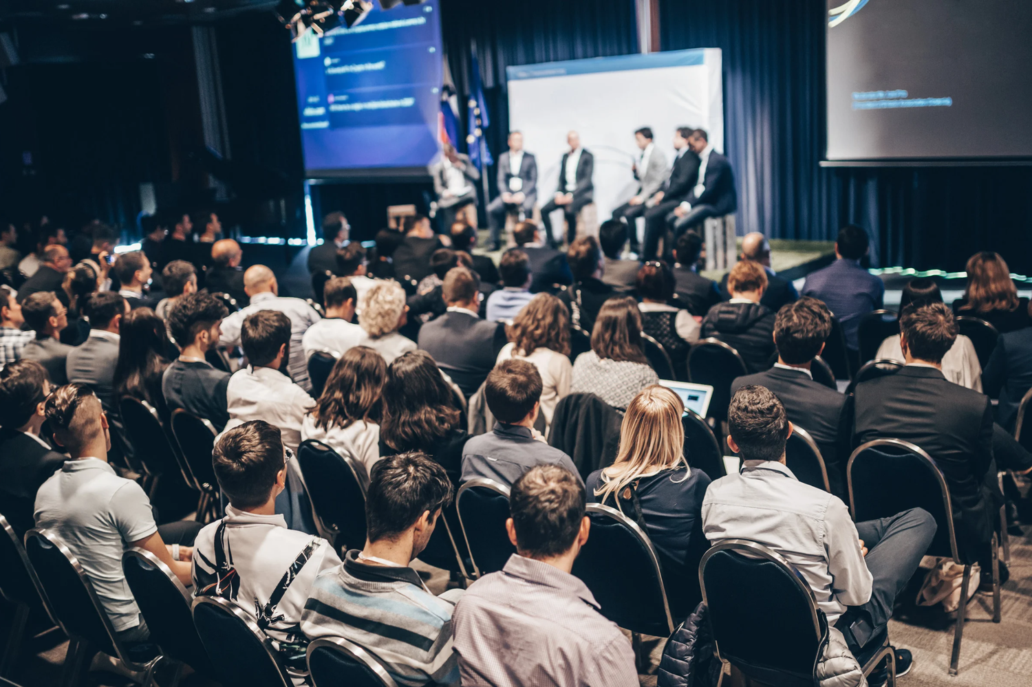 Large audience attending a professional business conference panel discussion in a darkened auditorium.