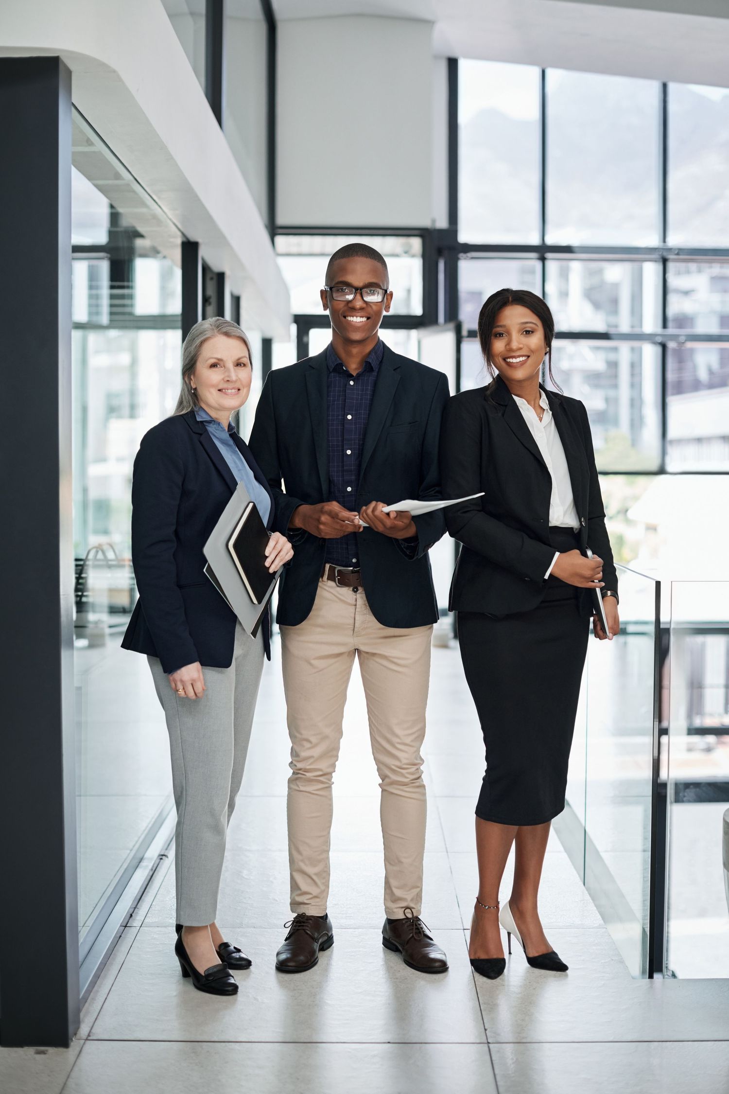Three diverse business professionals smiling together in a modern corporate office lobby.