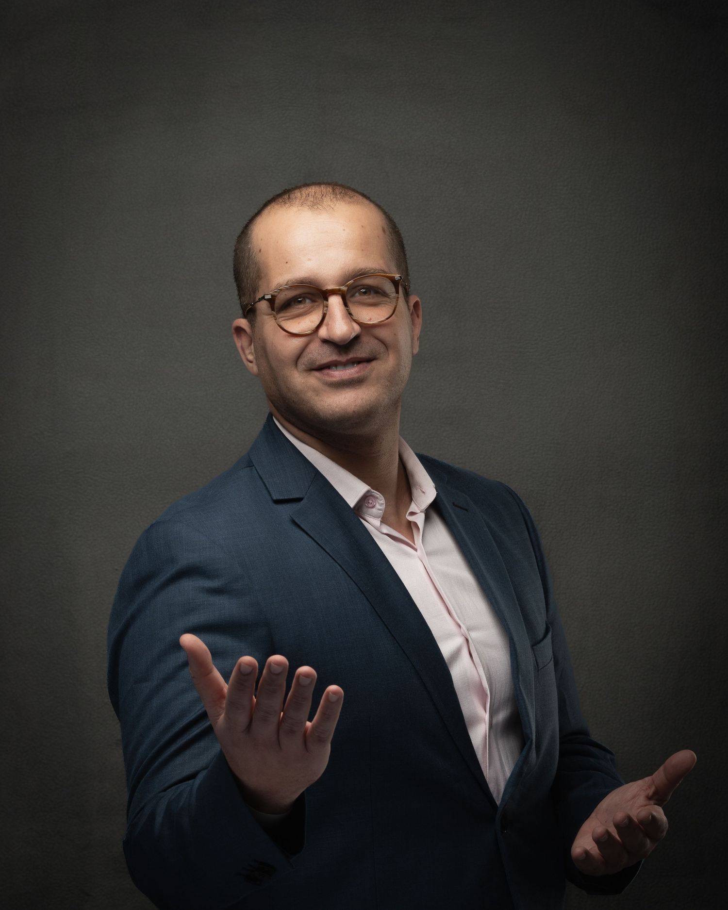 Smiling bald man in blue blazer wearing glasses gesturing with open hands against gray background.