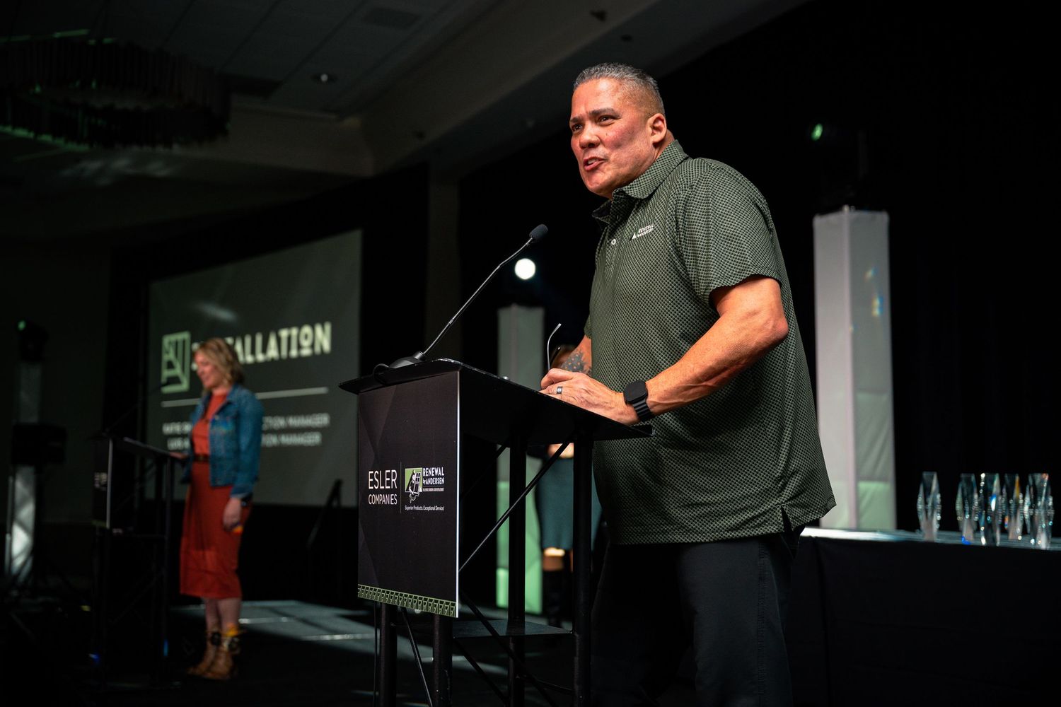 Man in green polo shirt speaks at podium during formal installation ceremony event in conference hall.