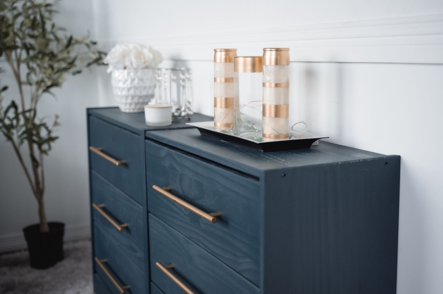 Navy blue dresser with copper handles styled with decor items and a potted plant in a modern bedroom setting.
