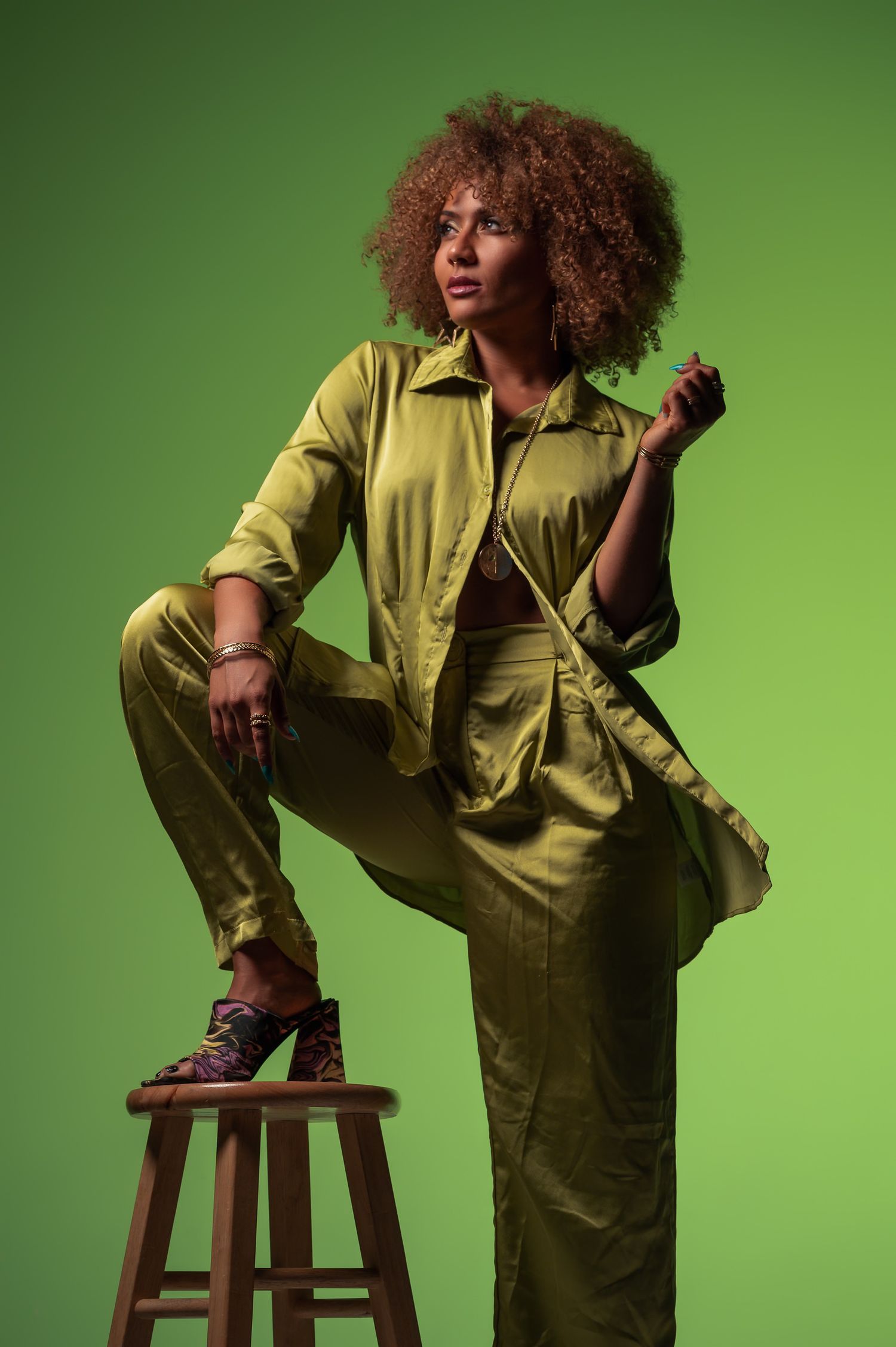 Woman in green satin suit with afro hairstyle posing on wooden stool against green background.