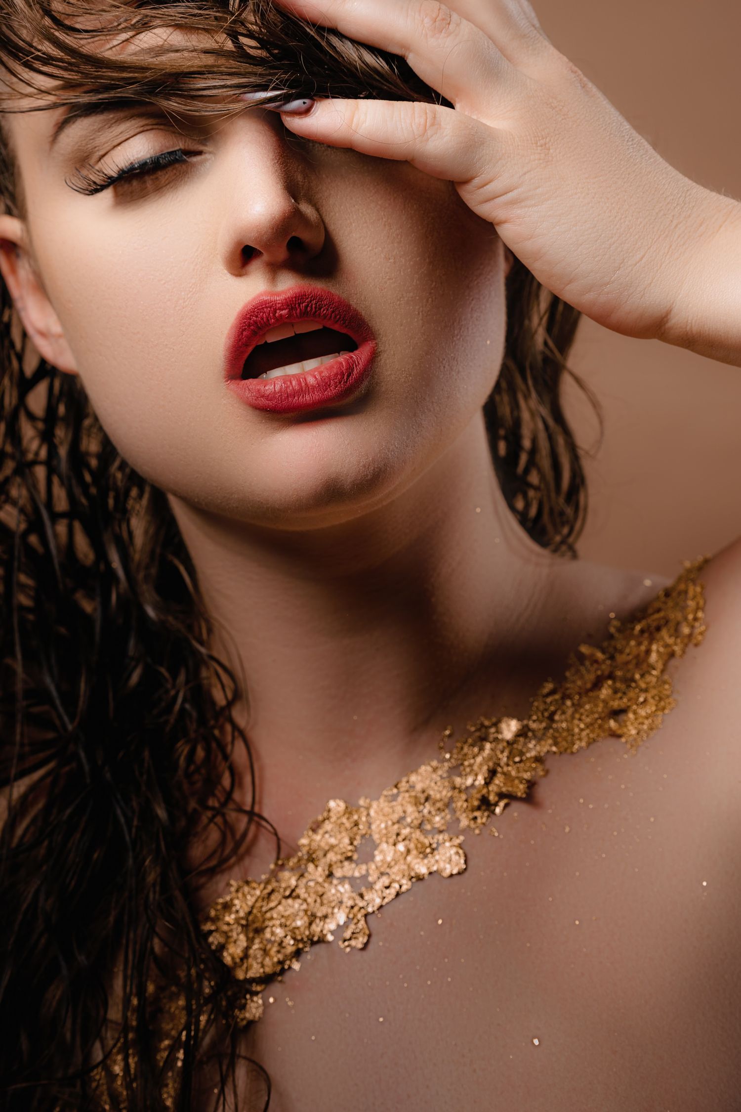 Woman with wet hair, red lips, and gold flakes on her chest poses dramatically against a beige background.