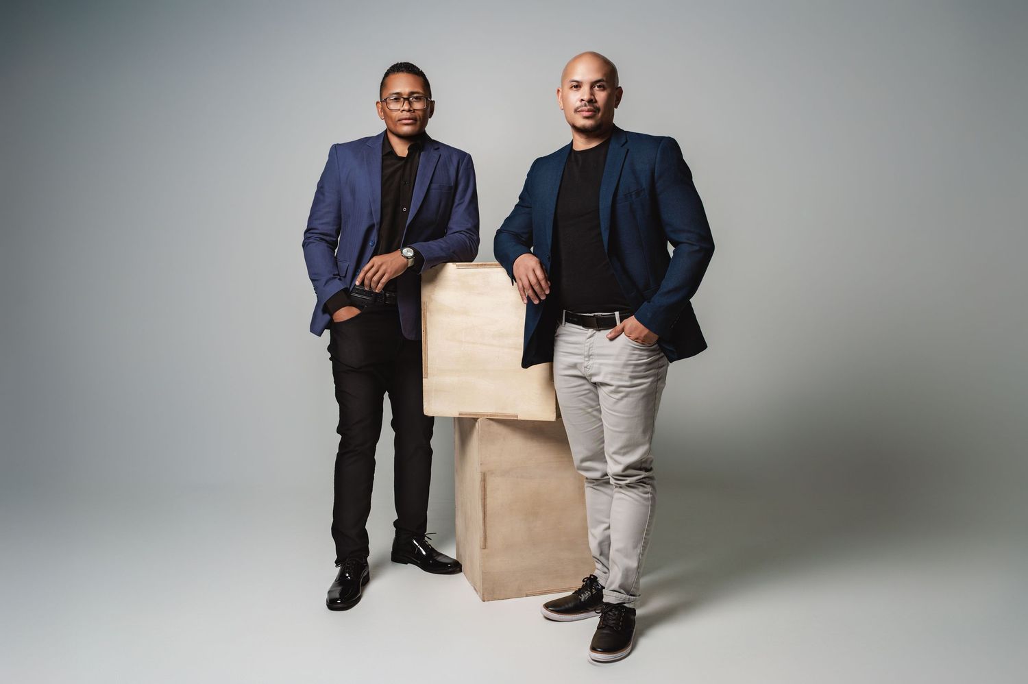 Professional studio photos show someone in a navy blazer and beige pants sitting on a modern cube ottoman against gray backdrop.