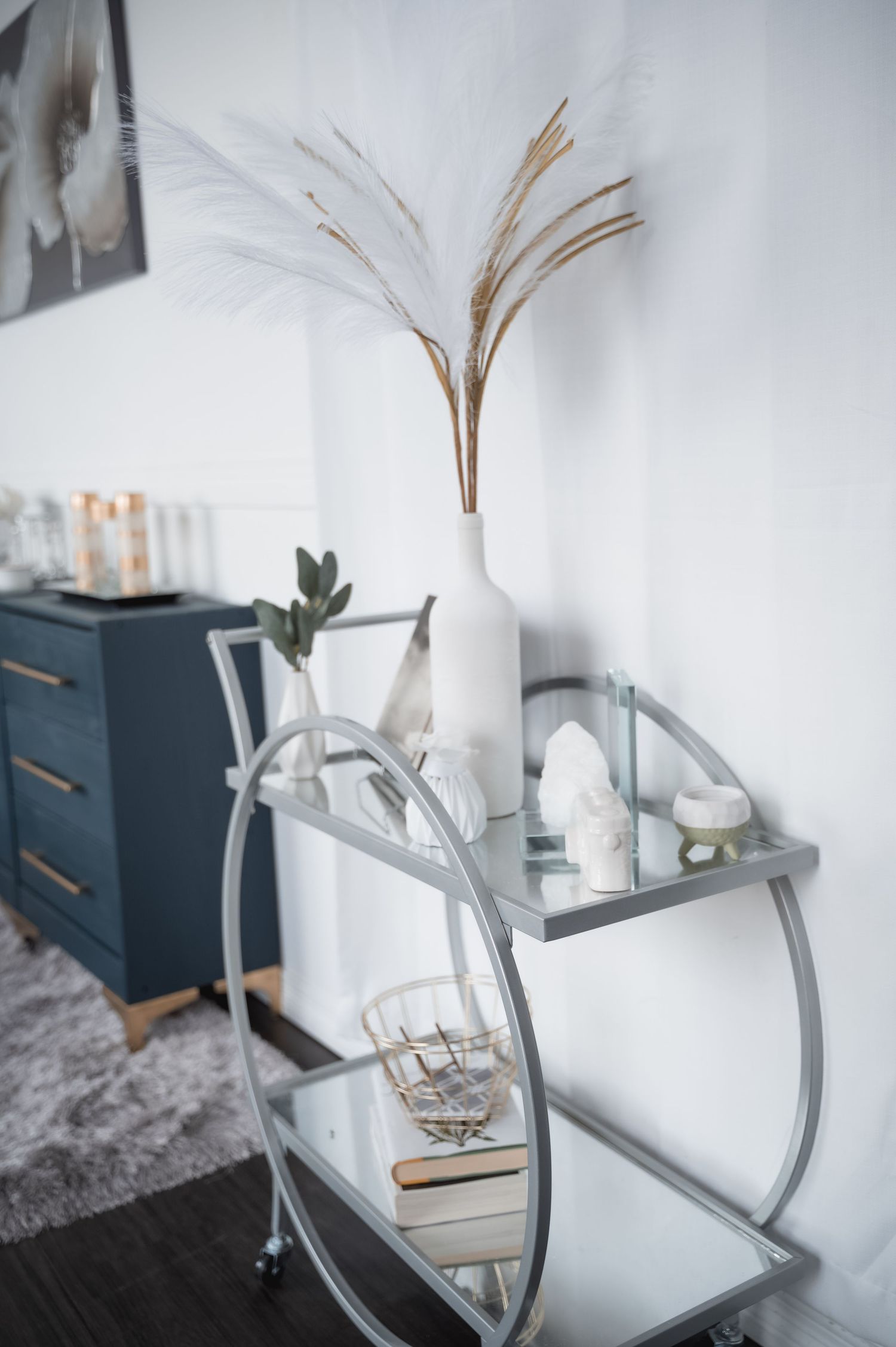 Modern white bedroom decor featuring a sleek navy dresser, glass vase with dried pampas grass, and minimalist accessories.