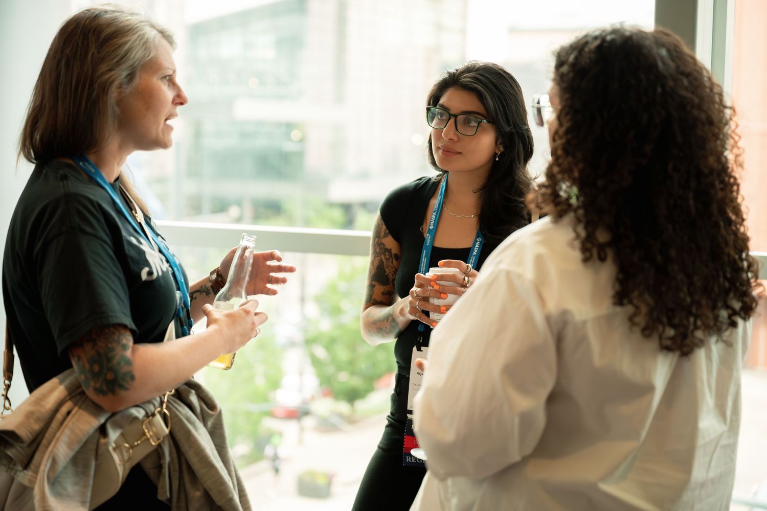 Three women engaged in conversation at a professional networking event, standing near large windows in a modern building.