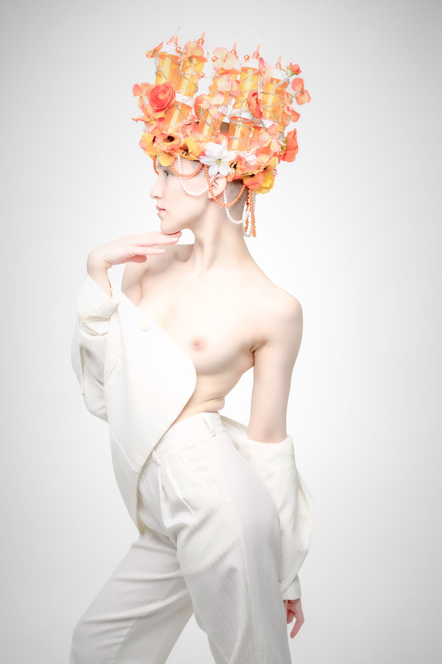 A model poses in white pants and jacket with an elaborate orange floral headdress against a light background.