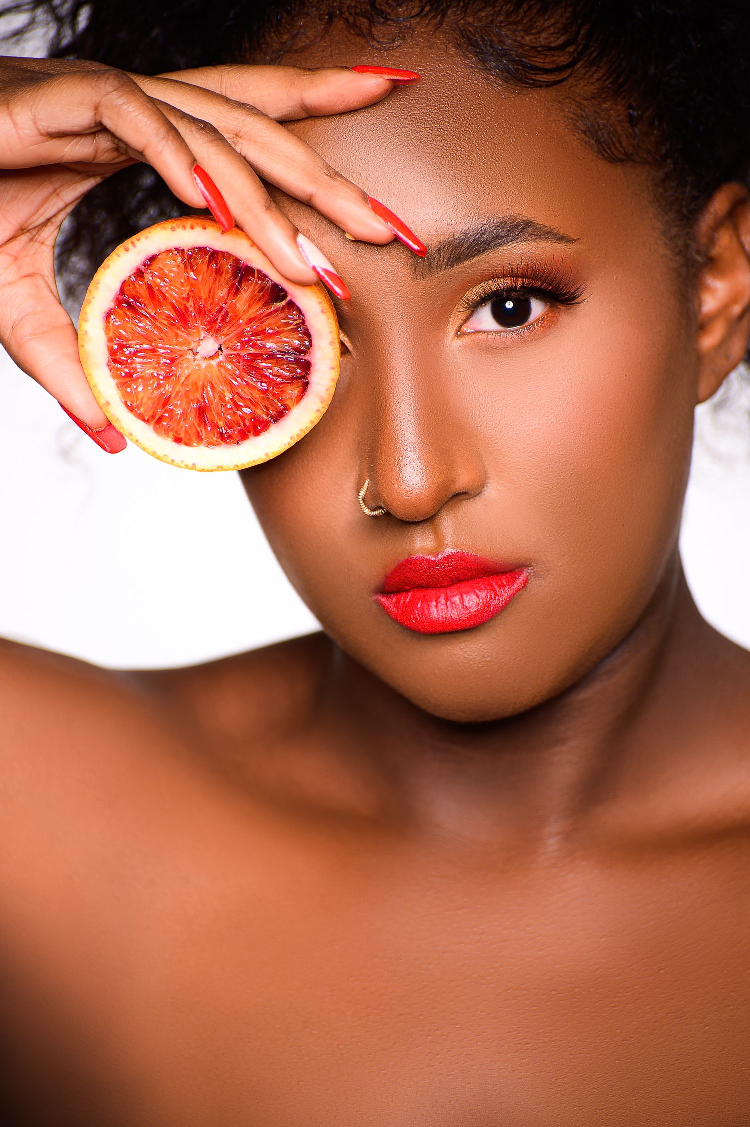 A young Black woman with red lips holds a sliced blood orange over one eye against a white background.