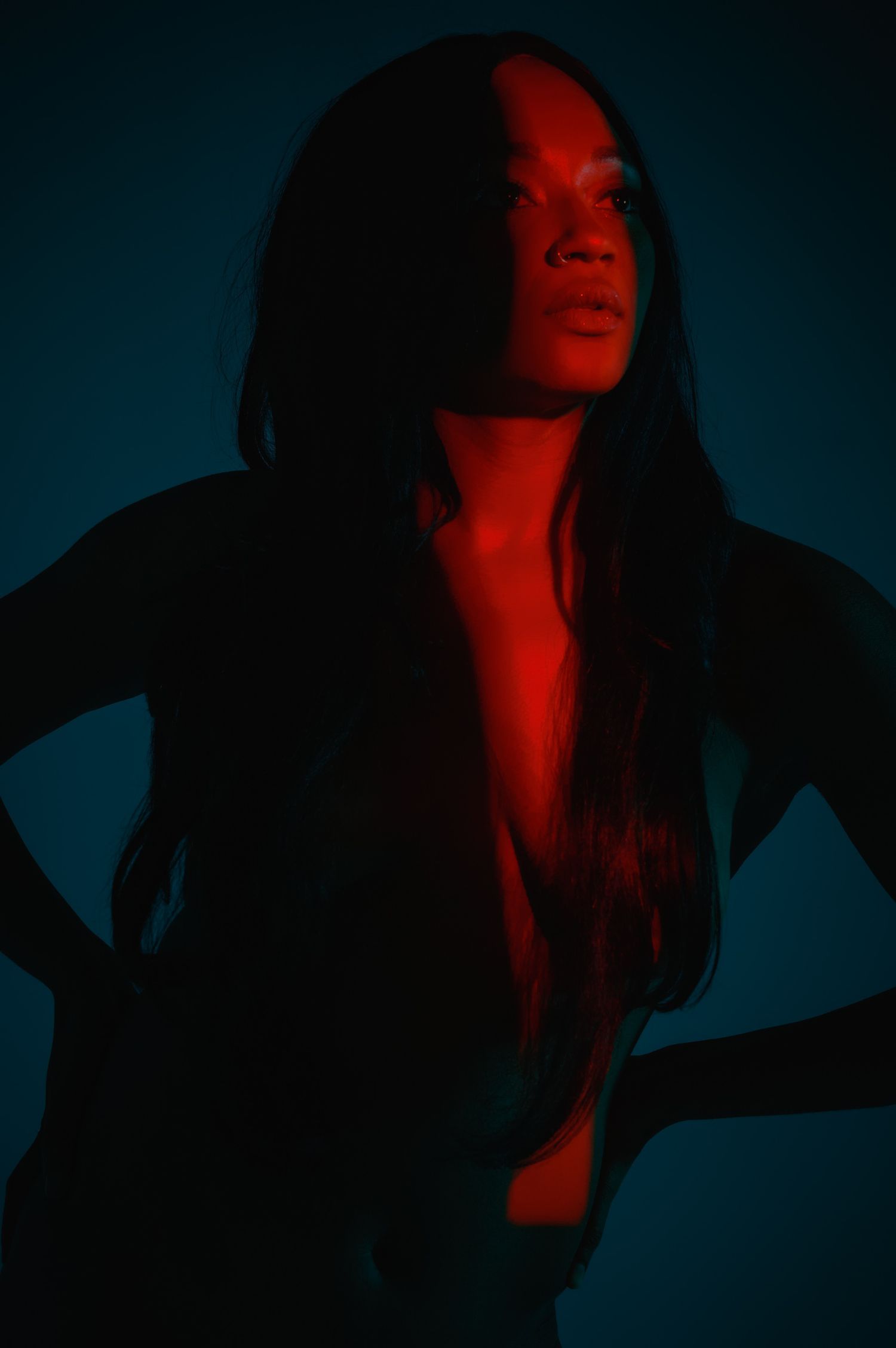 A woman with long dark hair posed dramatically under red and teal studio lighting in a moody portrait.