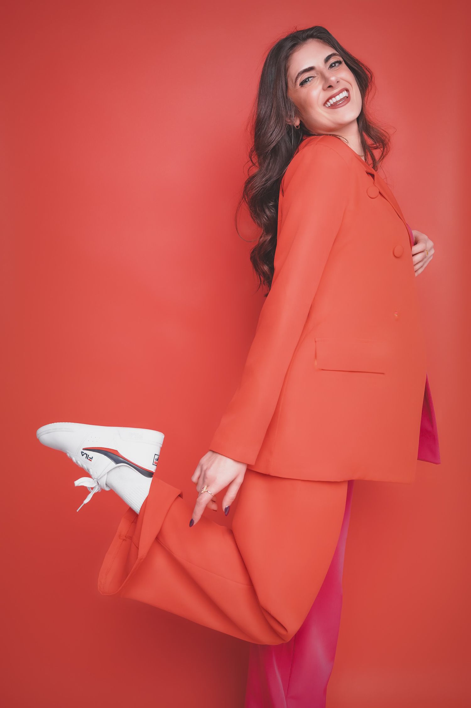 Woman in orange blazer and pink pants laughs playfully, holding a red bag against a vibrant red background.