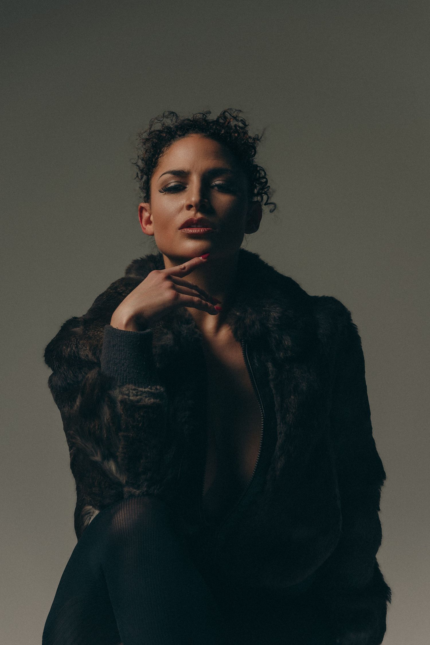 A woman in a dark fur coat poses confidently with hand under chin against a moody studio background.