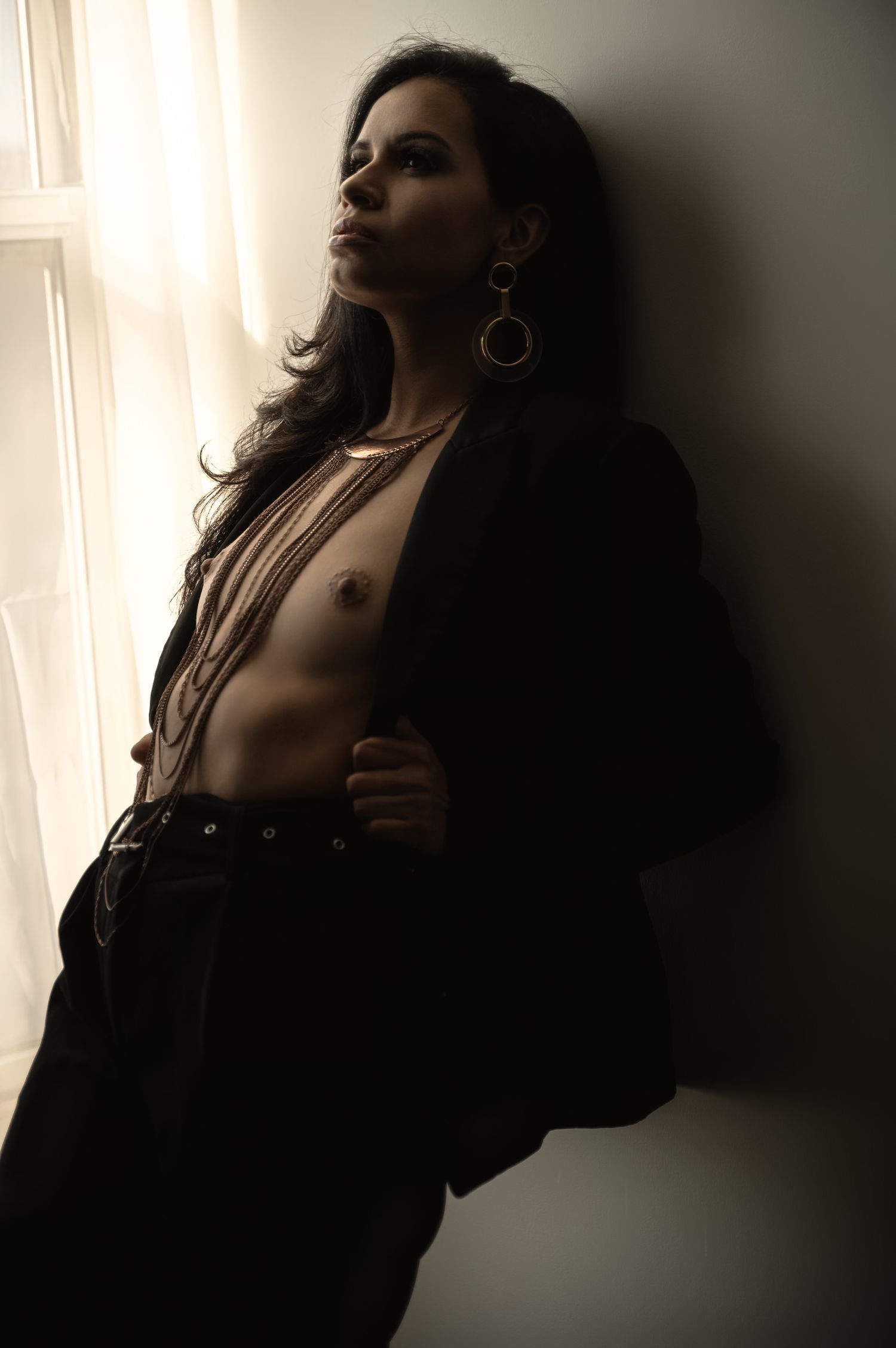 Dramatic low-key portrait of a woman in black blazer and jeans leaning against a wall by a bright window.
