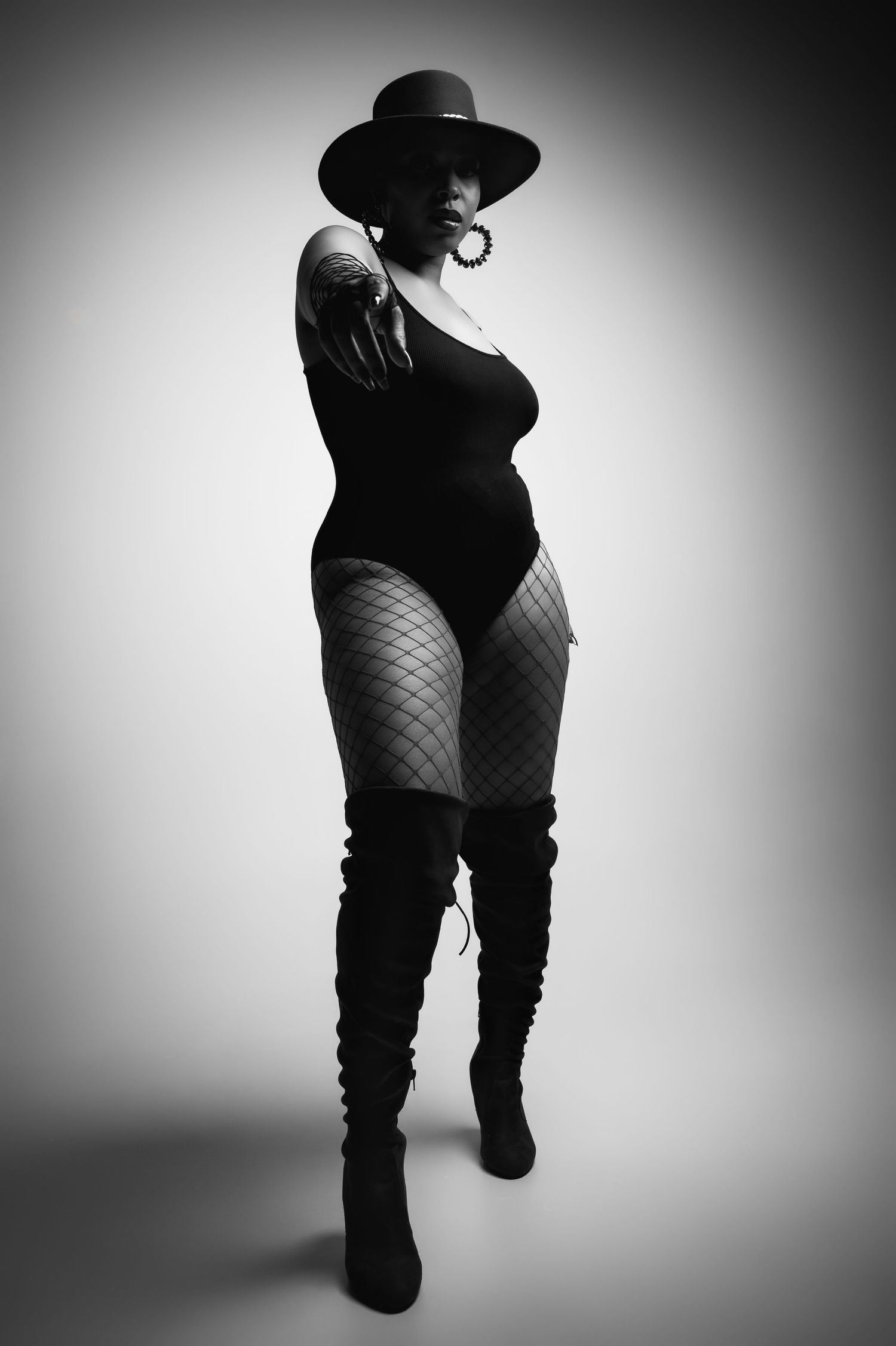 Dramatic black and white silhouette of a curvy figure in fishnet stockings, thigh-high boots and wide-brimmed hat.