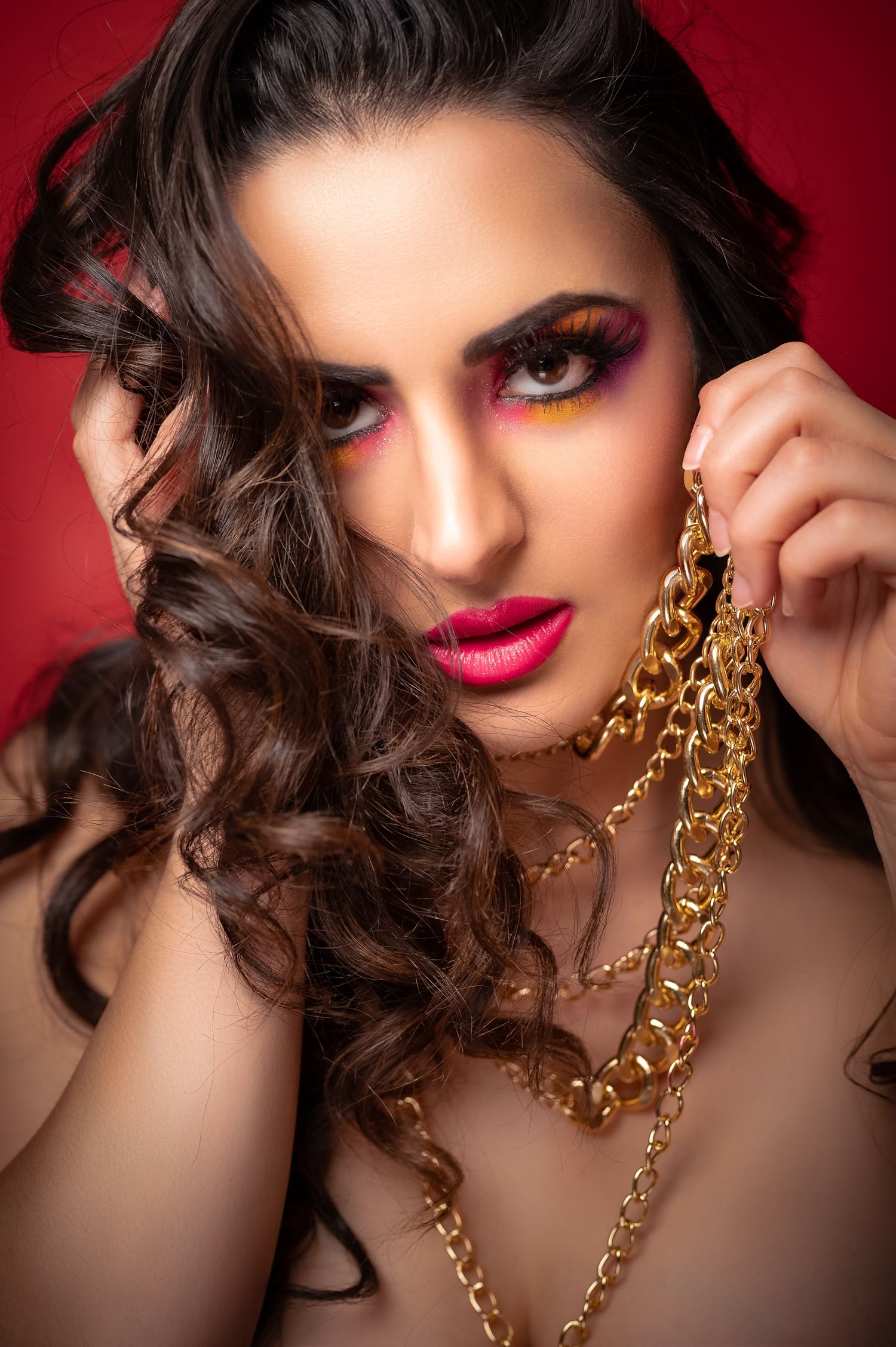 Woman with bold colorful eye makeup and red lips holding gold chain necklaces against a red background.
