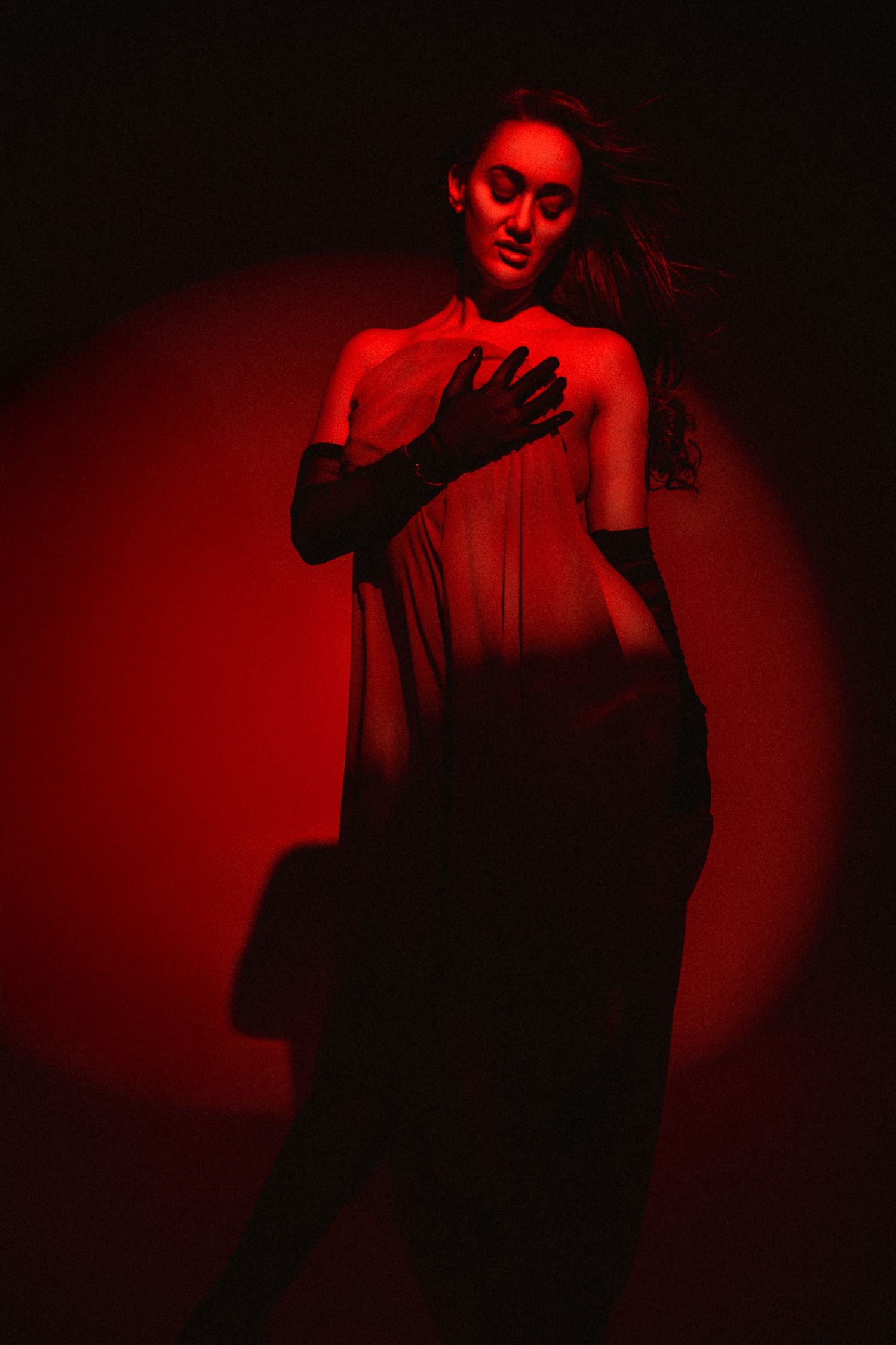 Woman in sheer dress and black gloves poses dramatically under red studio lighting.