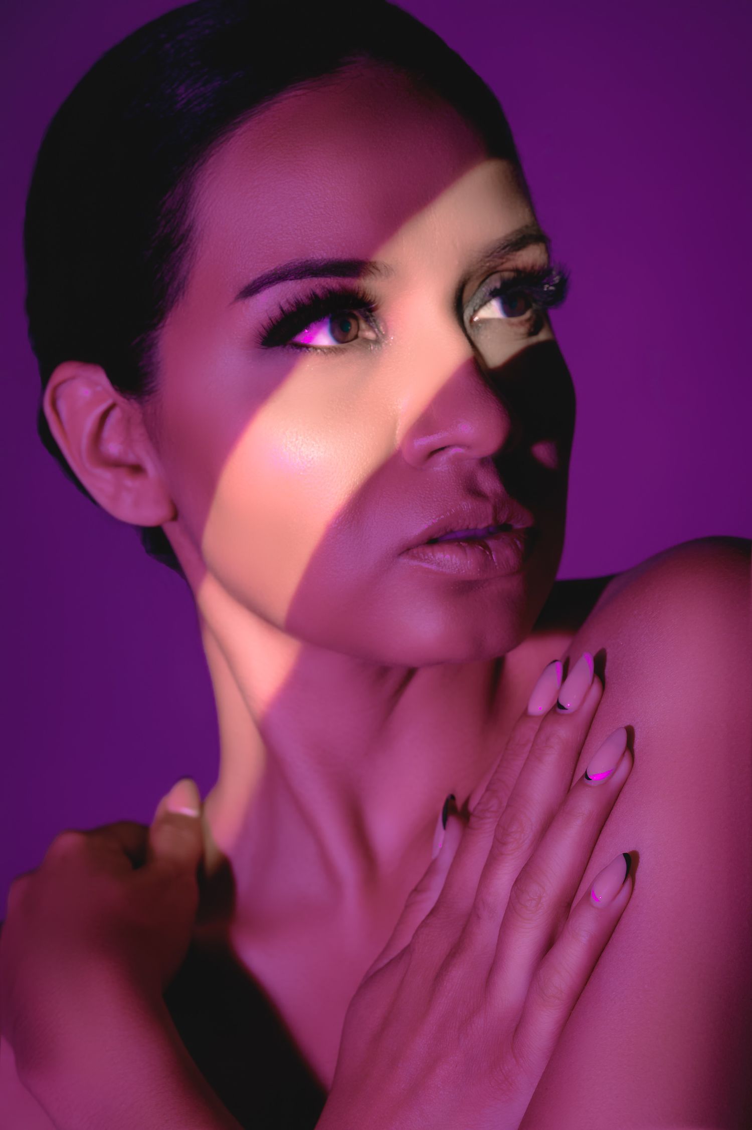 Woman with dramatic makeup and black nail tips posed against purple background with artistic light projection on face.