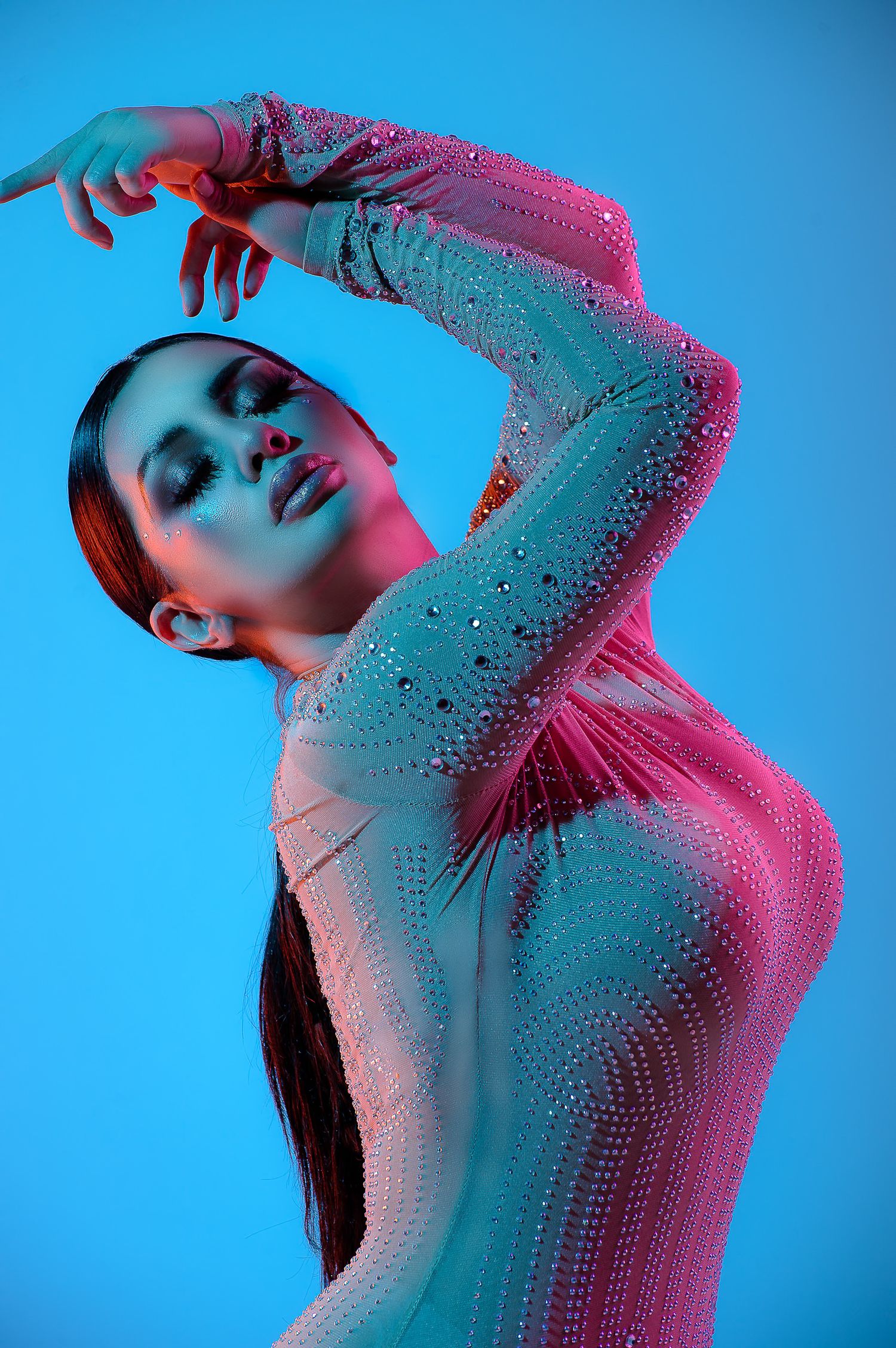 Woman in a sequined bodysuit poses gracefully under pink and blue neon lights against a blue background.
