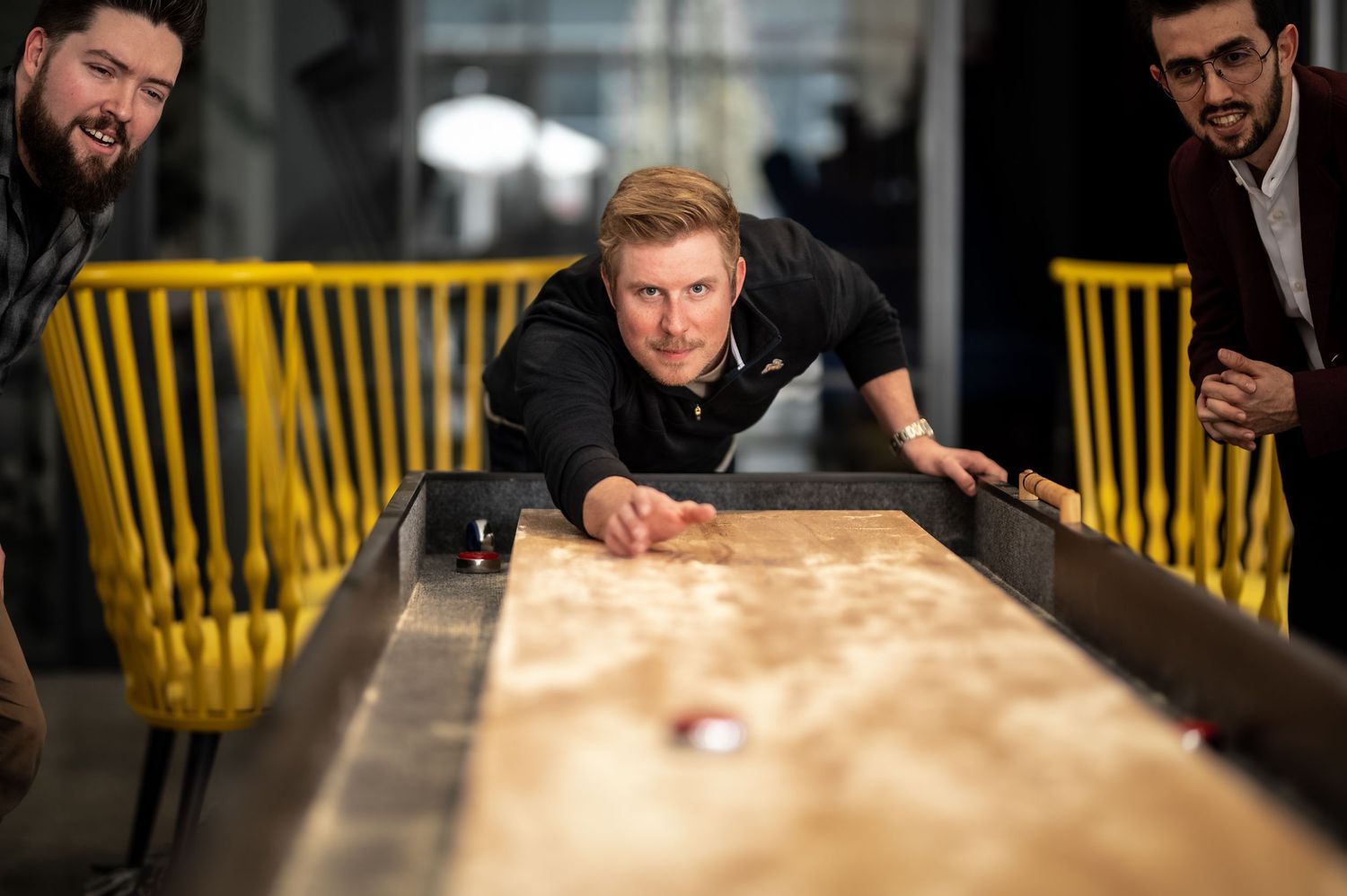 People playing shuffleboard at a bar with yellow chairs in the background while leaning over the wooden table.