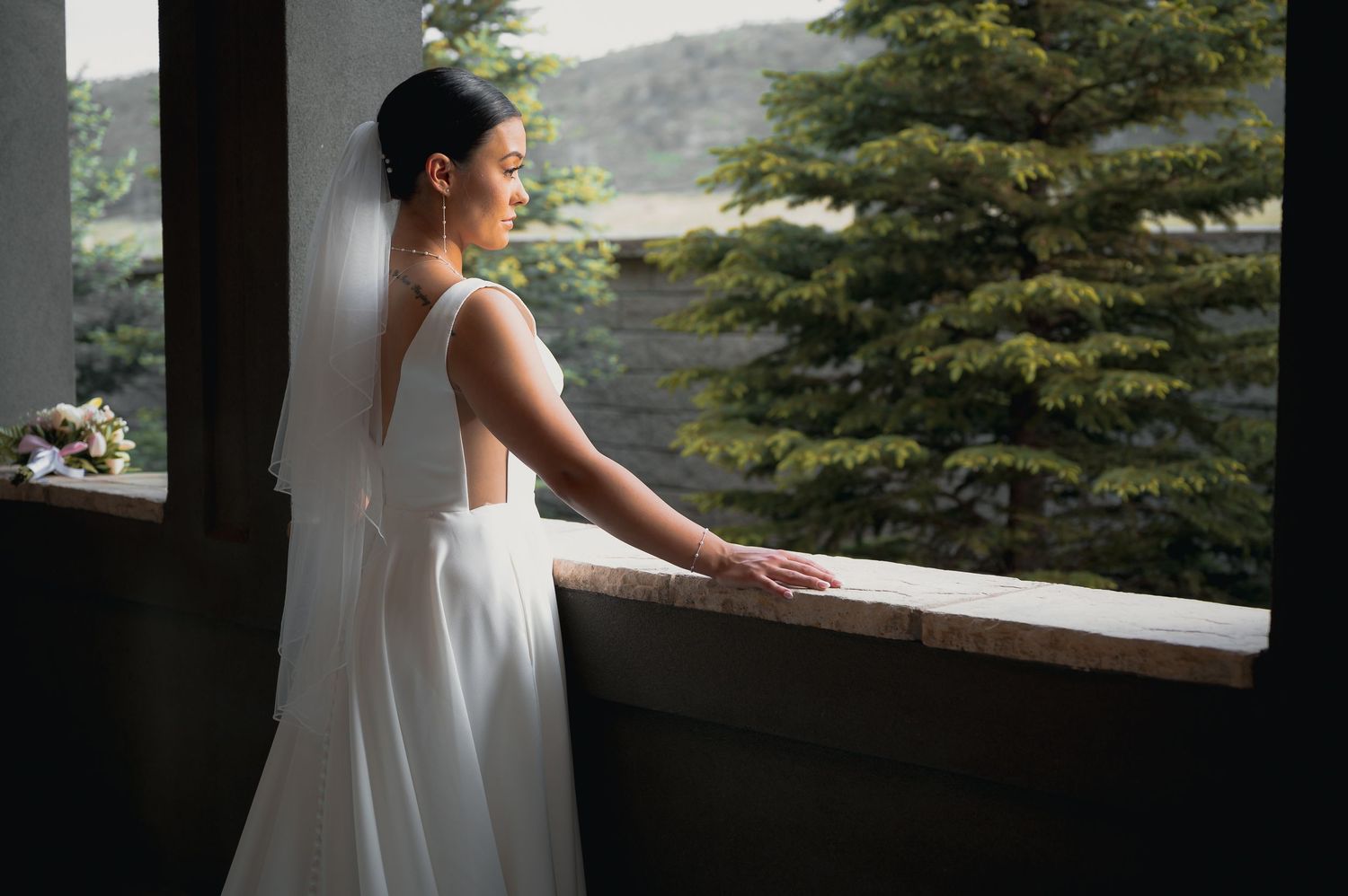 Bride in white gown and veil gazes out window at evergreen trees, elegant backless dress detail visible.