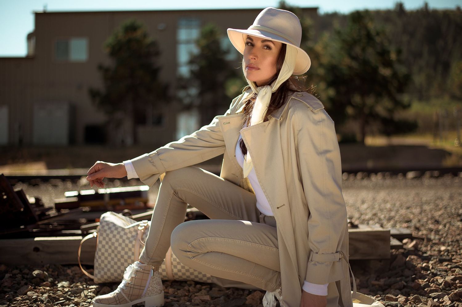 Woman in beige trench coat and white hat sitting on railroad tracks in an outdoor urban setting.