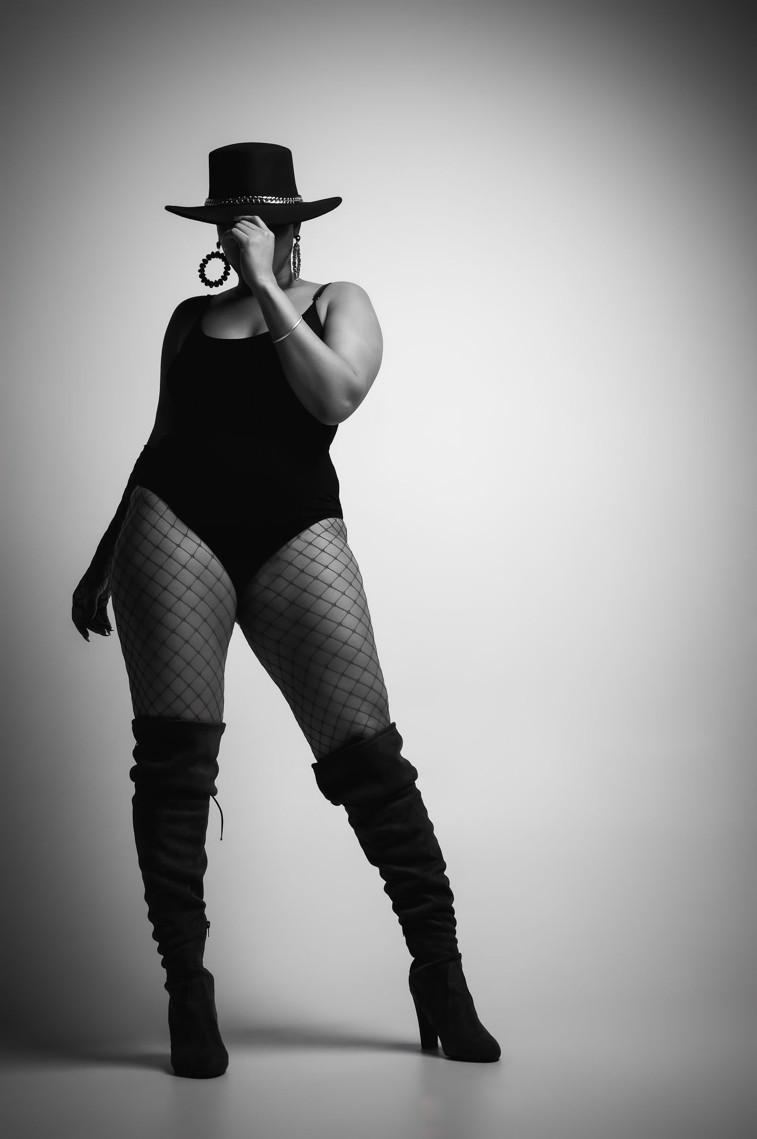 Black and white photo of a woman in fishnet tights, bodysuit, thigh-high boots, and wide-brim hat posing confidently.