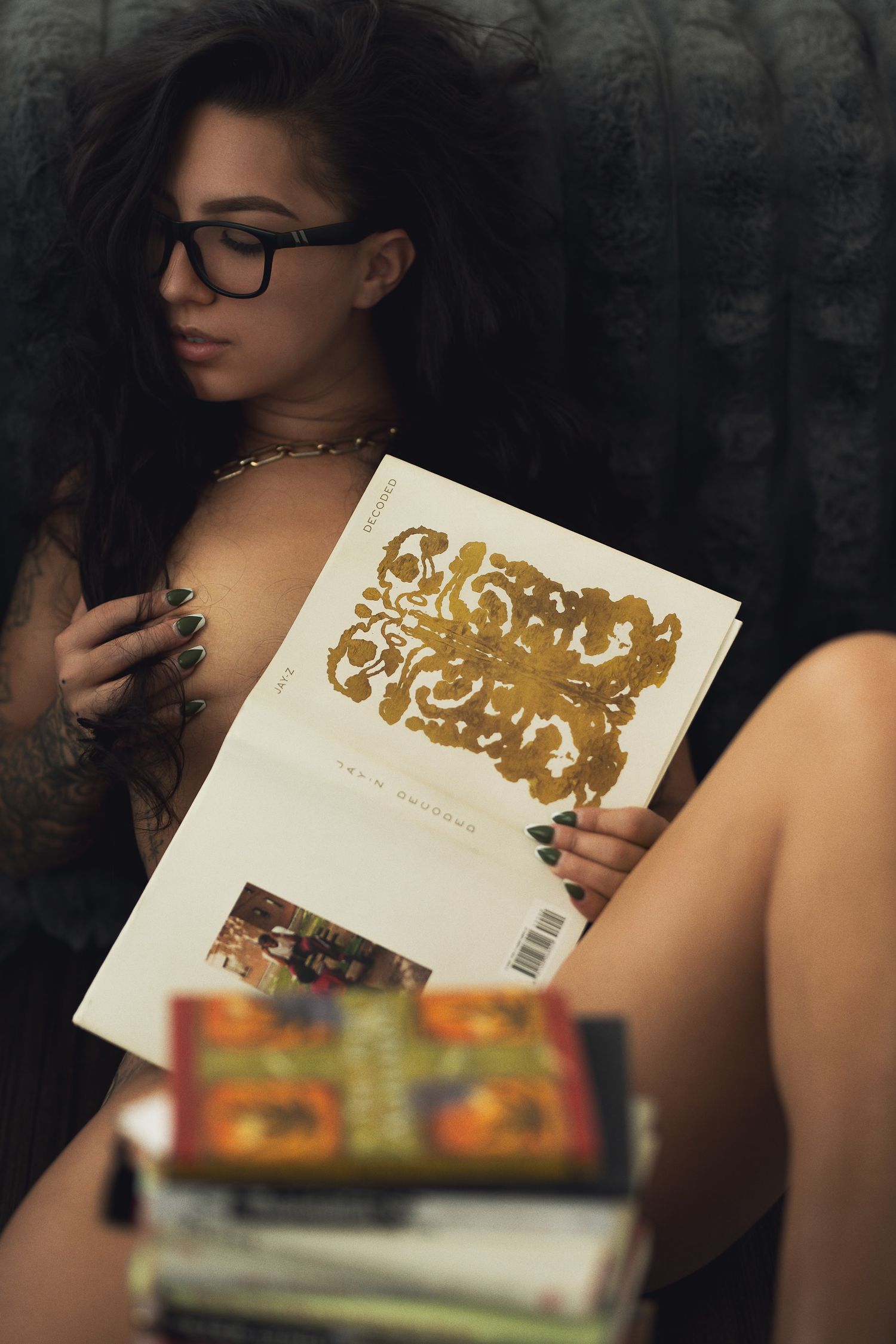 Young woman with glasses and tattoos reading a gold-covered book surrounded by a stack of colorful books.