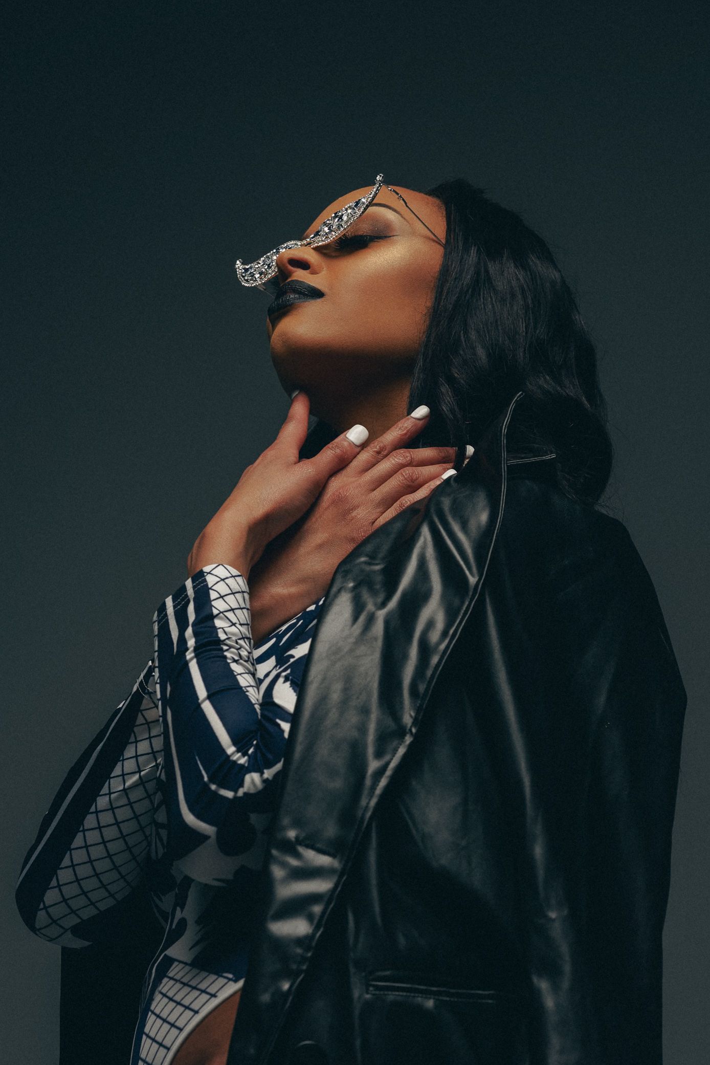 A woman in a black leather jacket and futuristic crystal sunglasses poses dramatically against a dark gray background.