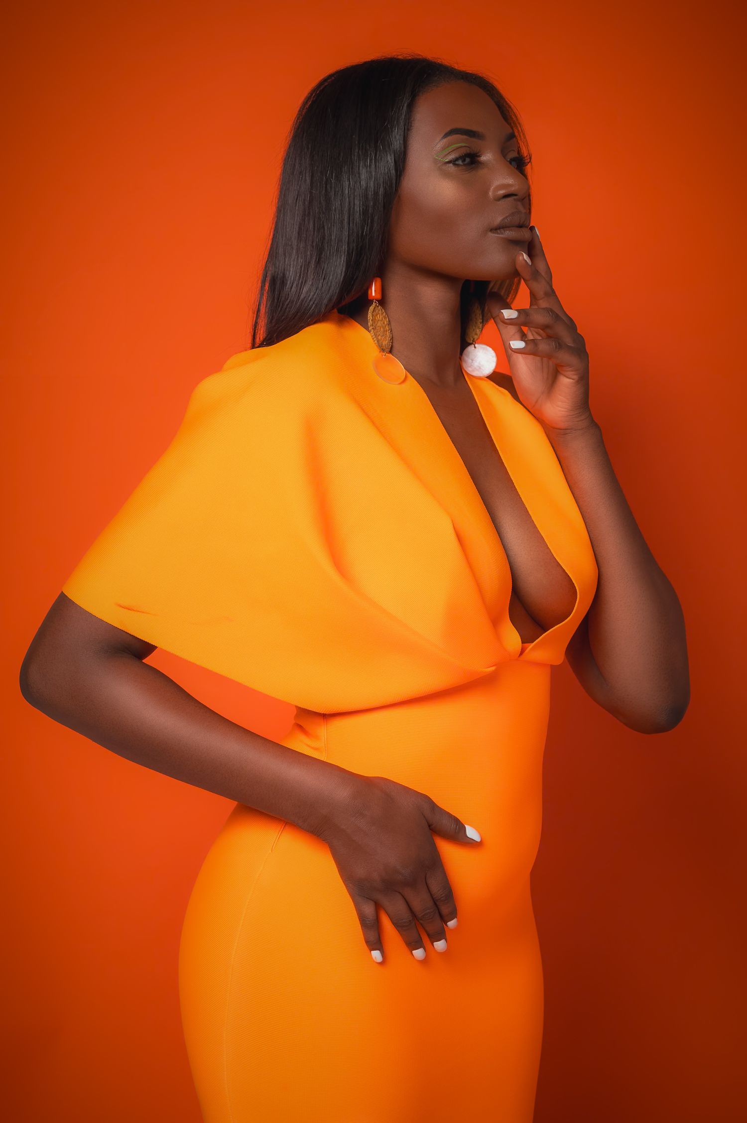 A woman in a vibrant orange dress poses confidently against an orange background, wearing large white earrings.