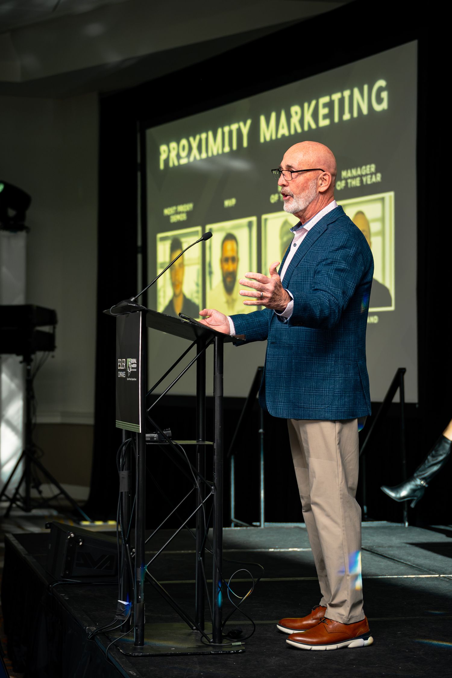 A speaker presents at a Proximity Marketing awards event, standing at a podium in a blue blazer before a large screen.