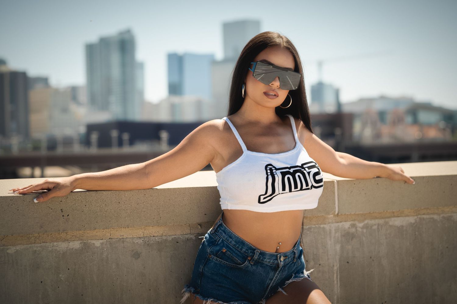 Athletic woman in Nike crop top and denim shorts poses against urban skyline backdrop on sunny day.
