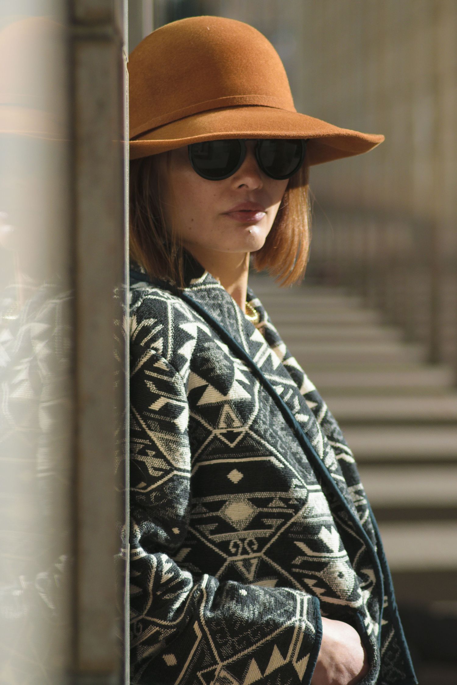 Person wearing a brown wide-brim hat and geometric patterned sweater poses against a blurred architectural background.