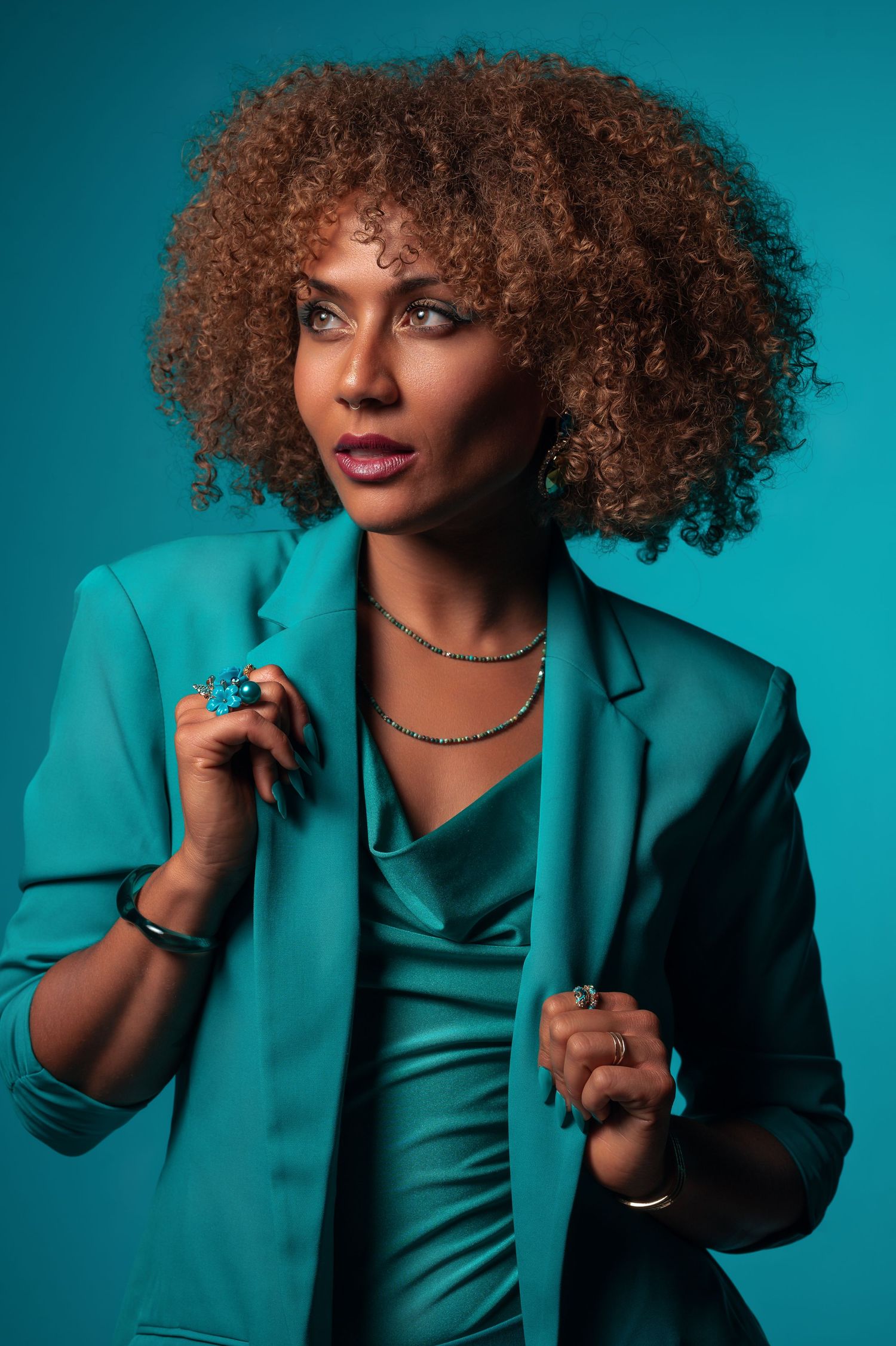 Fashion model in turquoise blazer poses expressively against blue backdrop with natural curly hair and statement jewelry.
