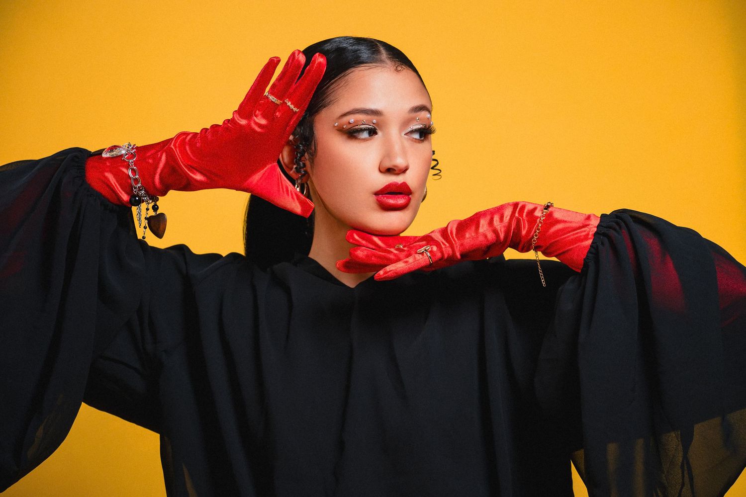 Woman in black outfit with red gloves and bold red lips poses against a vibrant yellow background.