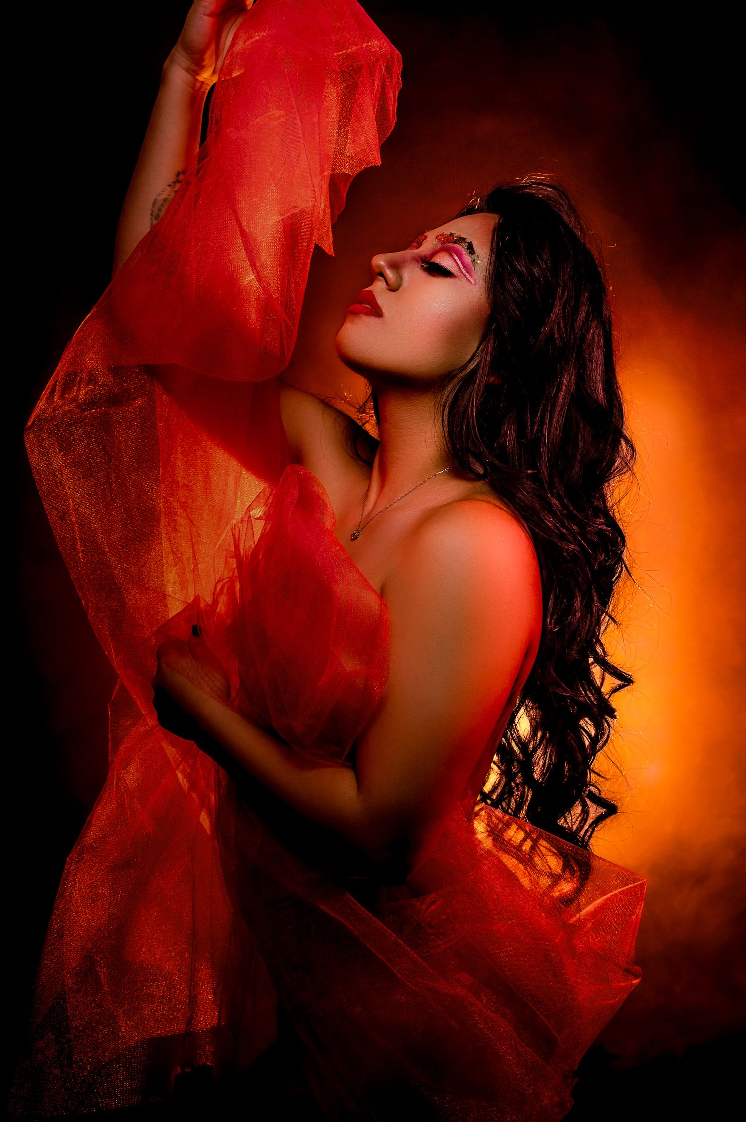 A woman in red draped fabric poses dramatically against a fiery orange backdrop with bold makeup and flowing dark hair.