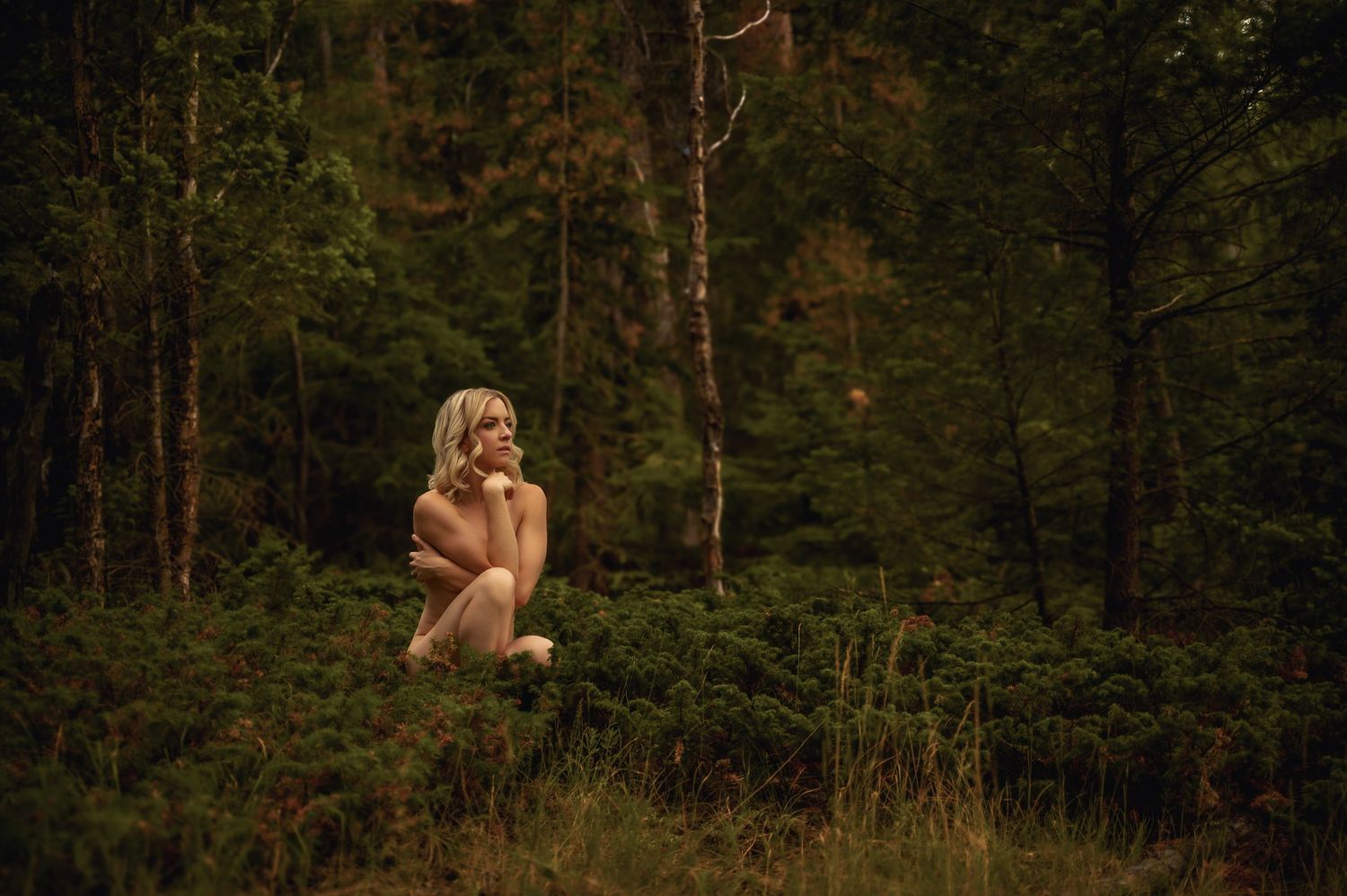 A blonde woman sits peacefully in a lush green forest clearing, surrounded by tall trees and dense foliage.