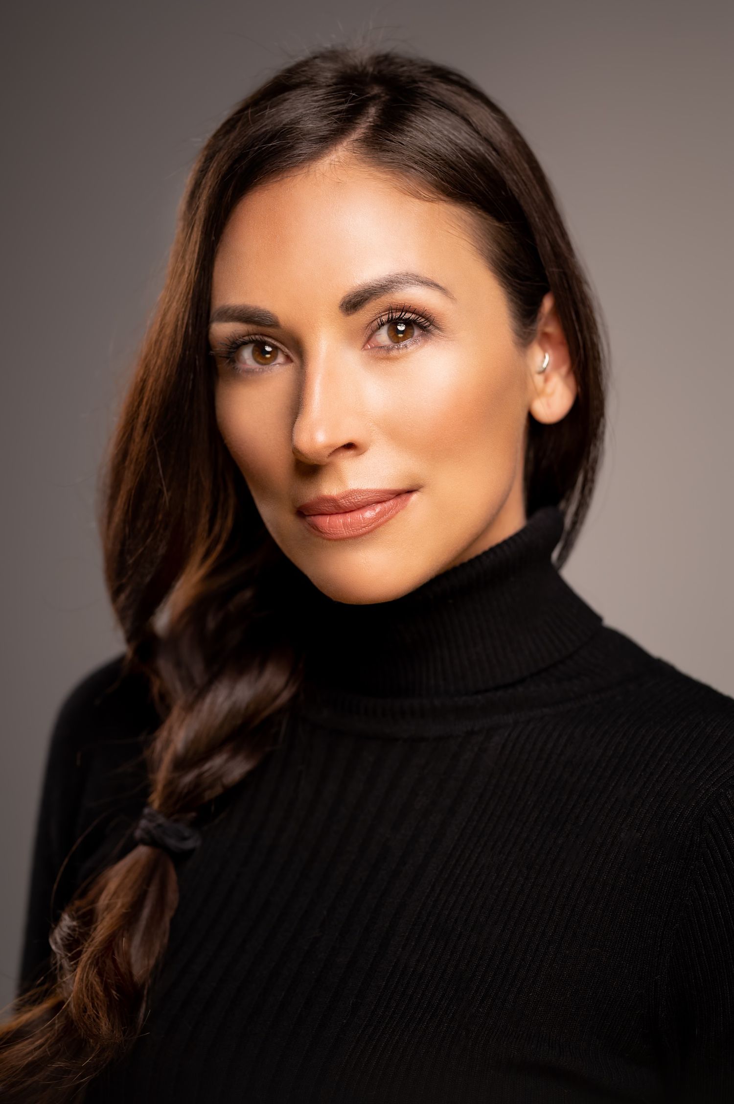Professional headshot of a brunette woman in a black turtleneck sweater posing against a gray background.