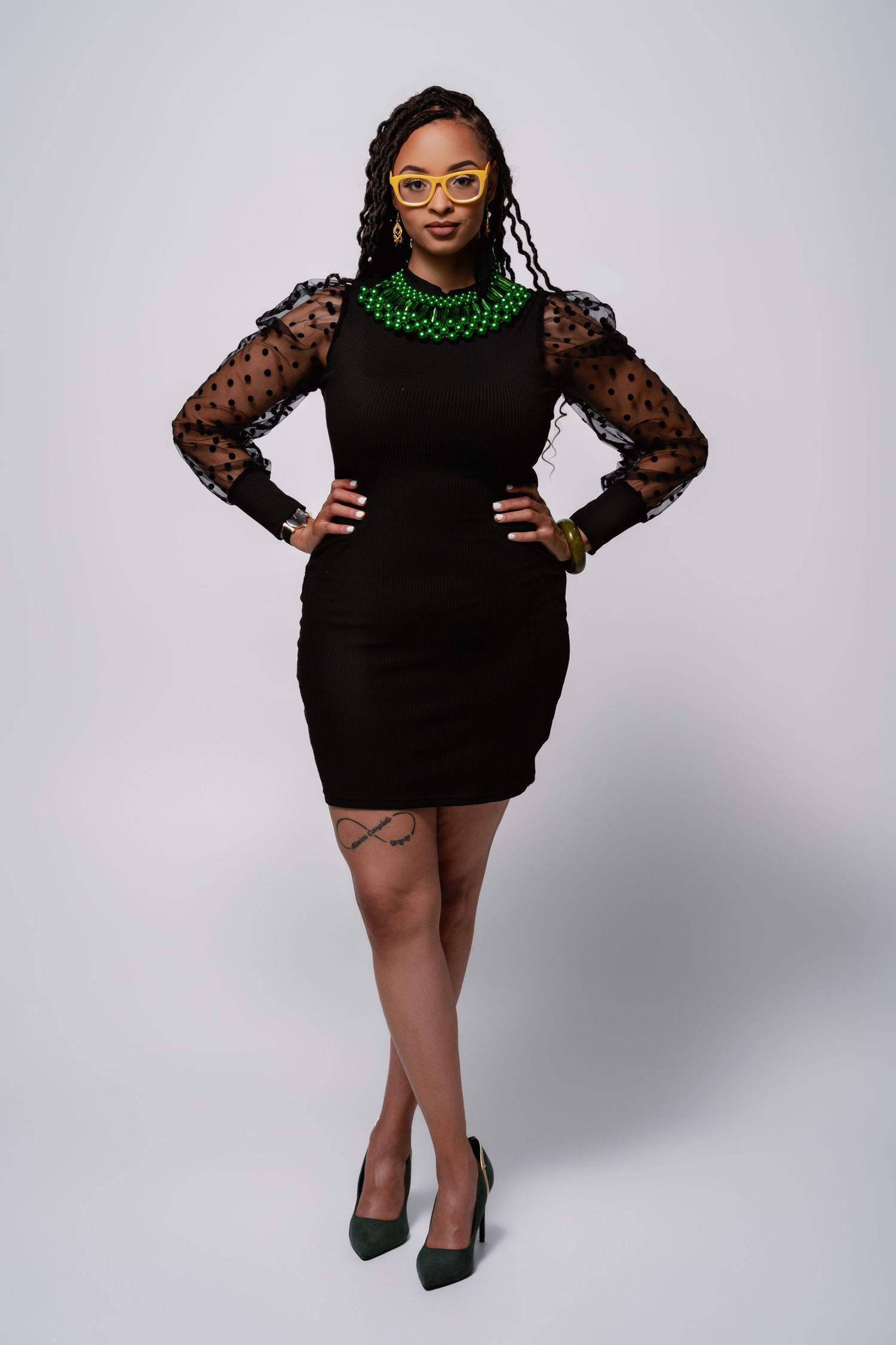 A fashion model poses in a black polka dot mesh sleeve dress with green accessories against a white backdrop.