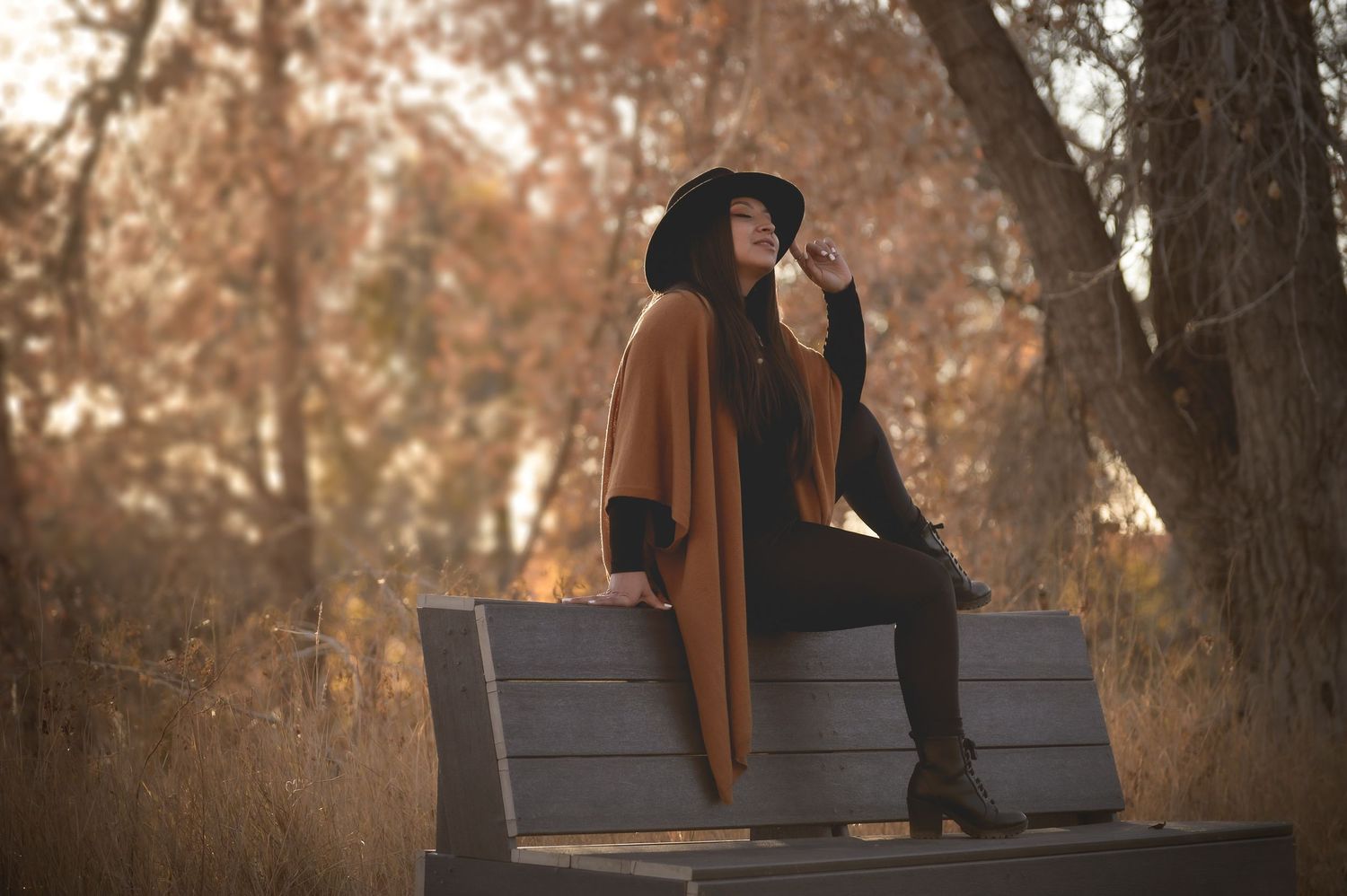 Woman in brown coat and black hat sitting on park bench surrounded by autumn trees.